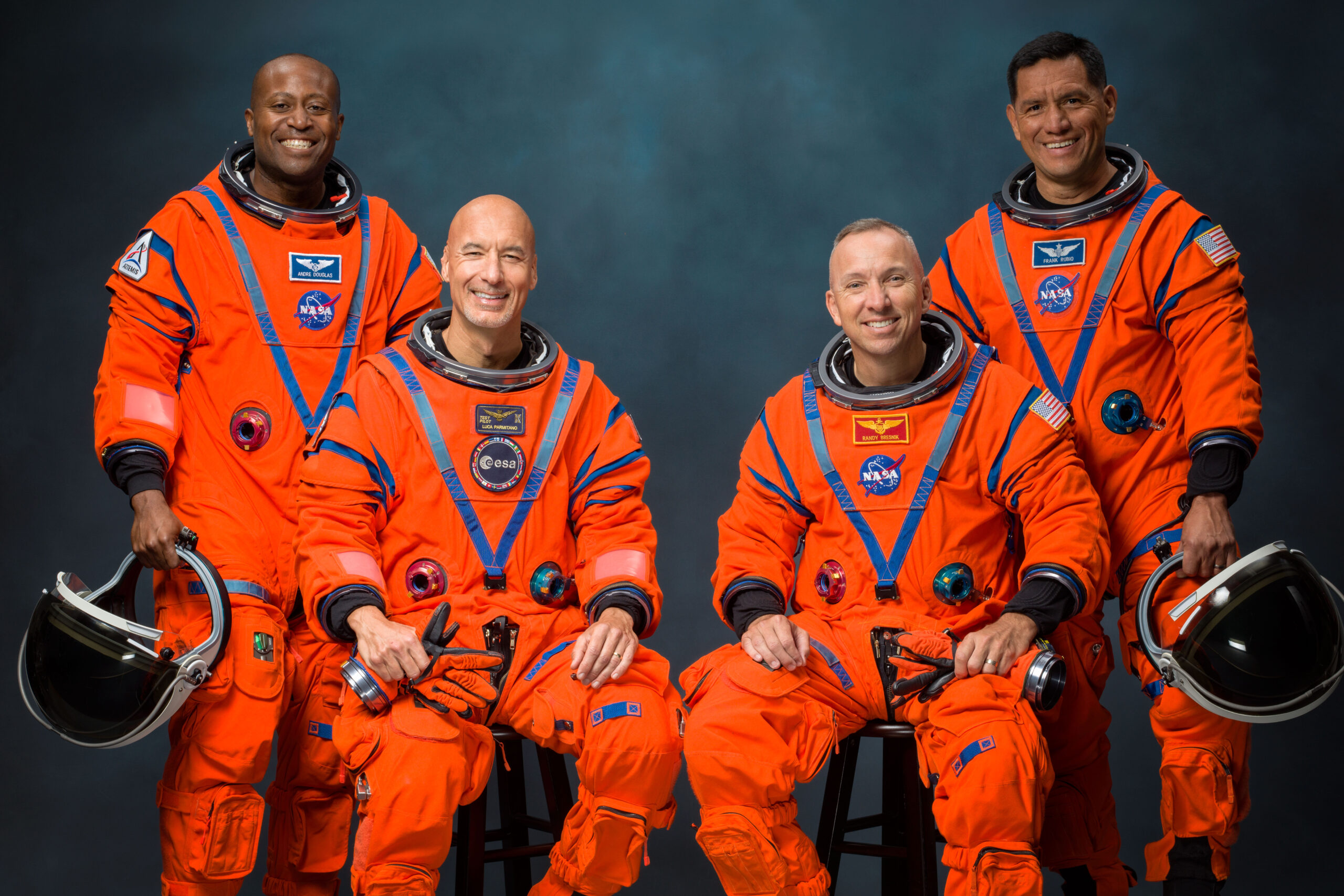 Four people in orange NASA suits pose together, holding helmets, against a dark neutral background.