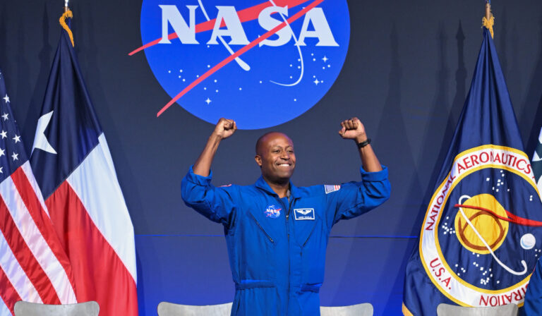 A person in a blue NASA spacesuit raises both arms before NASA logos and American flags.