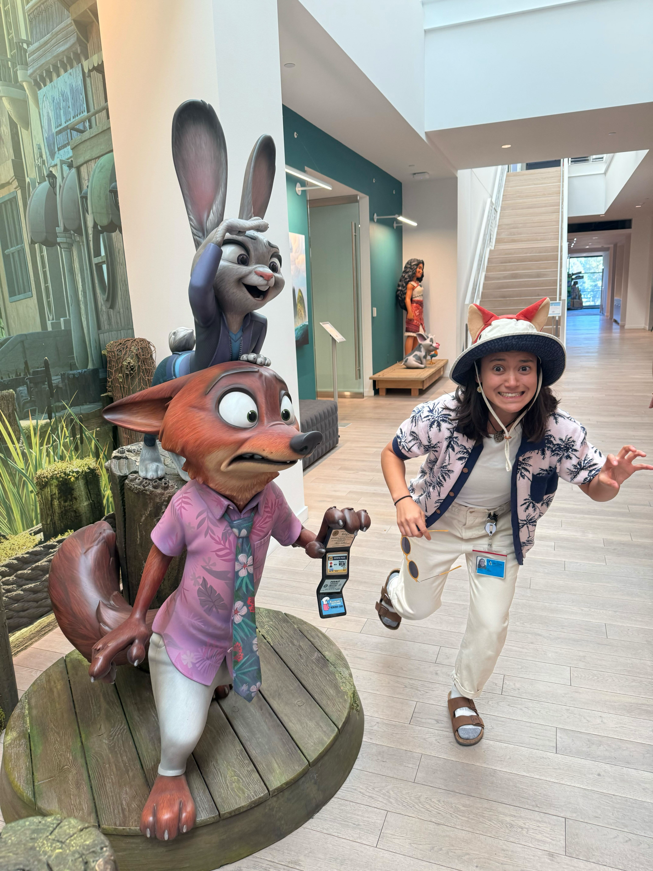 Amy, in a mostly white outfit with a hat, poses like she's running next to a statue of characters from Zootopia 2.