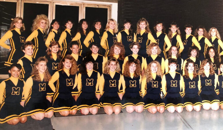 A large group of people in matching blue and yellow cheerleading uniforms pose indoors in front of large windows.