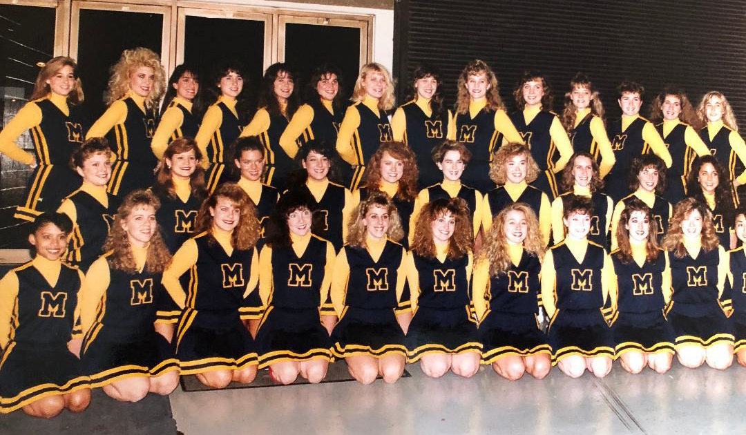 A large group of people in matching blue and yellow cheerleading uniforms pose indoors in front of large windows.