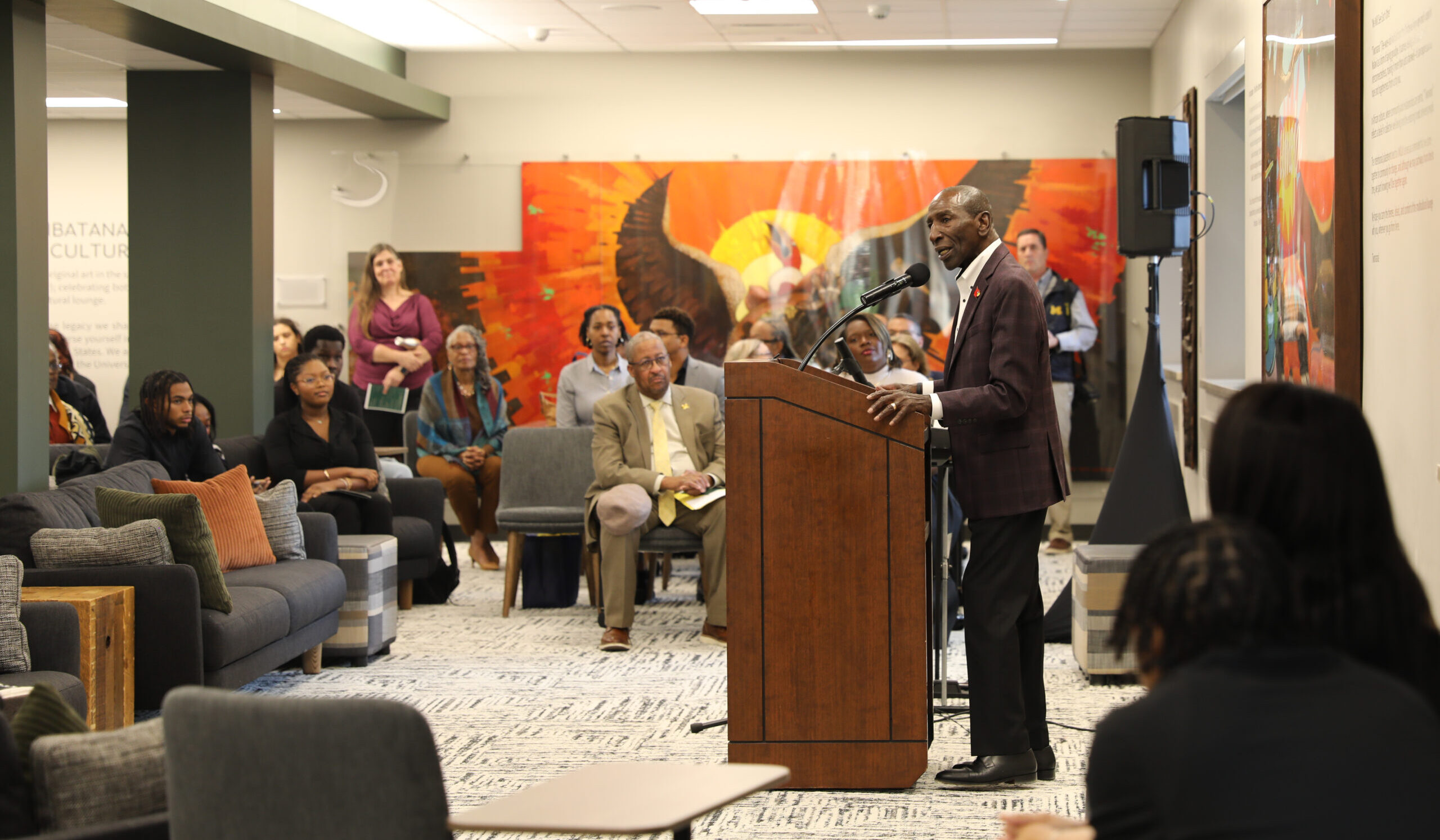 Lee Gill, in a dark suit, stands at a podium with people in seats listening. There is bright orange art on the wall.