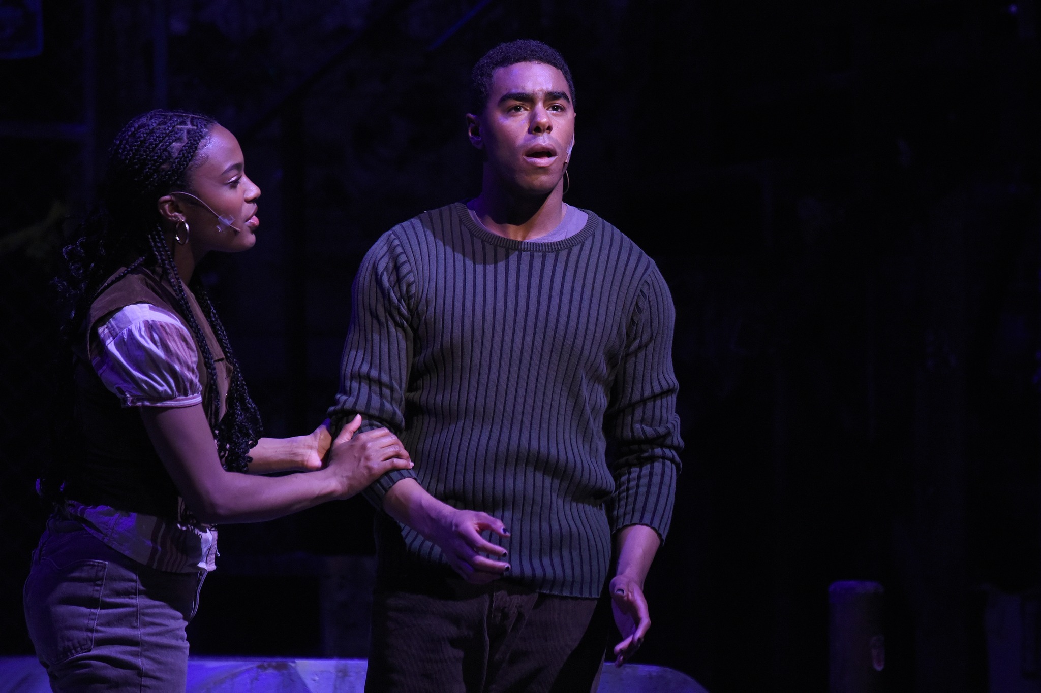 A dark photo of two students performing on stage. The one on the left is a woman wearing a short sleeve shirt and on the right is a man in a dark sweater.