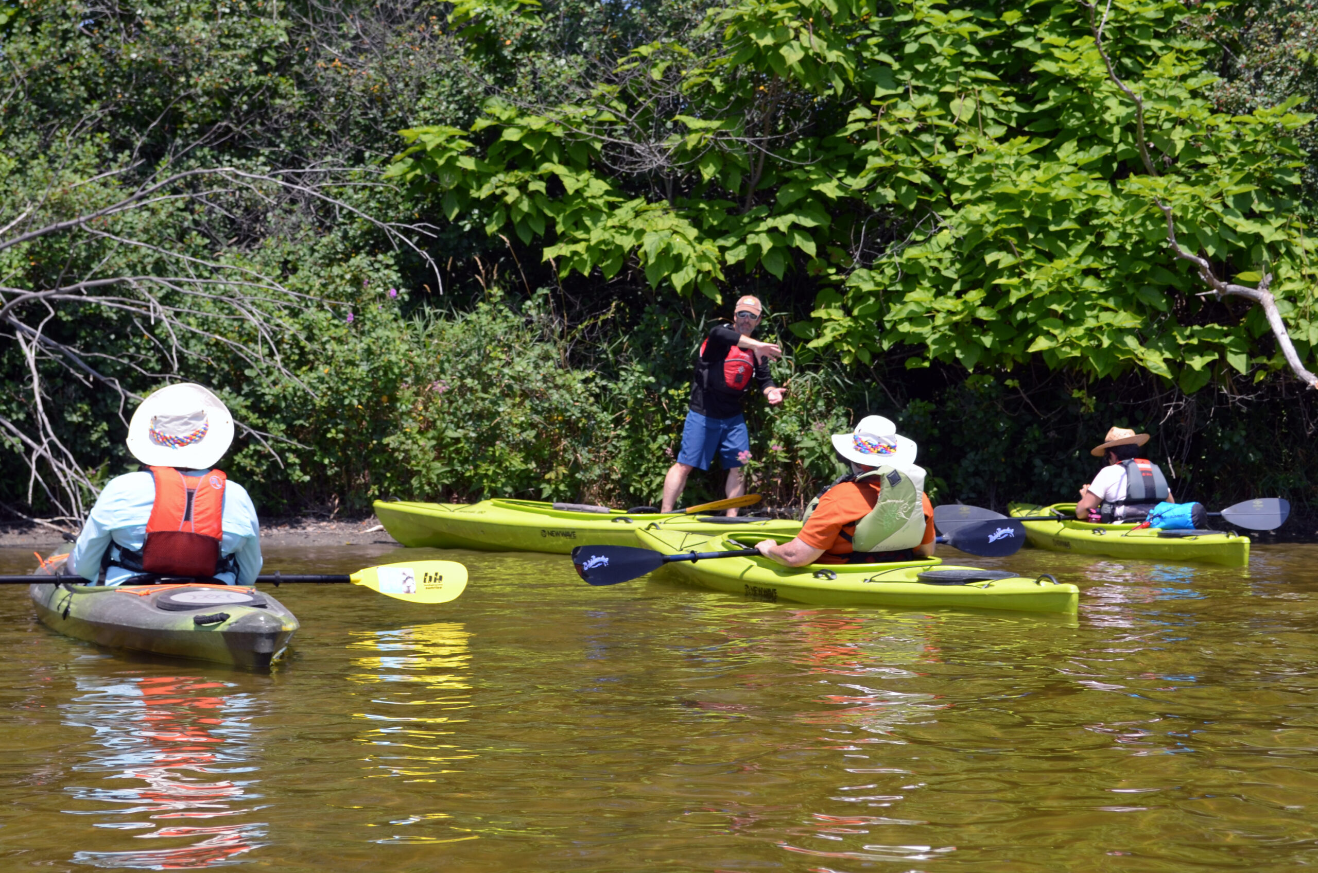 People in kayaks listen to a person standing out of their kayak, talking. The kayaks are on shallow water with tress in the background.