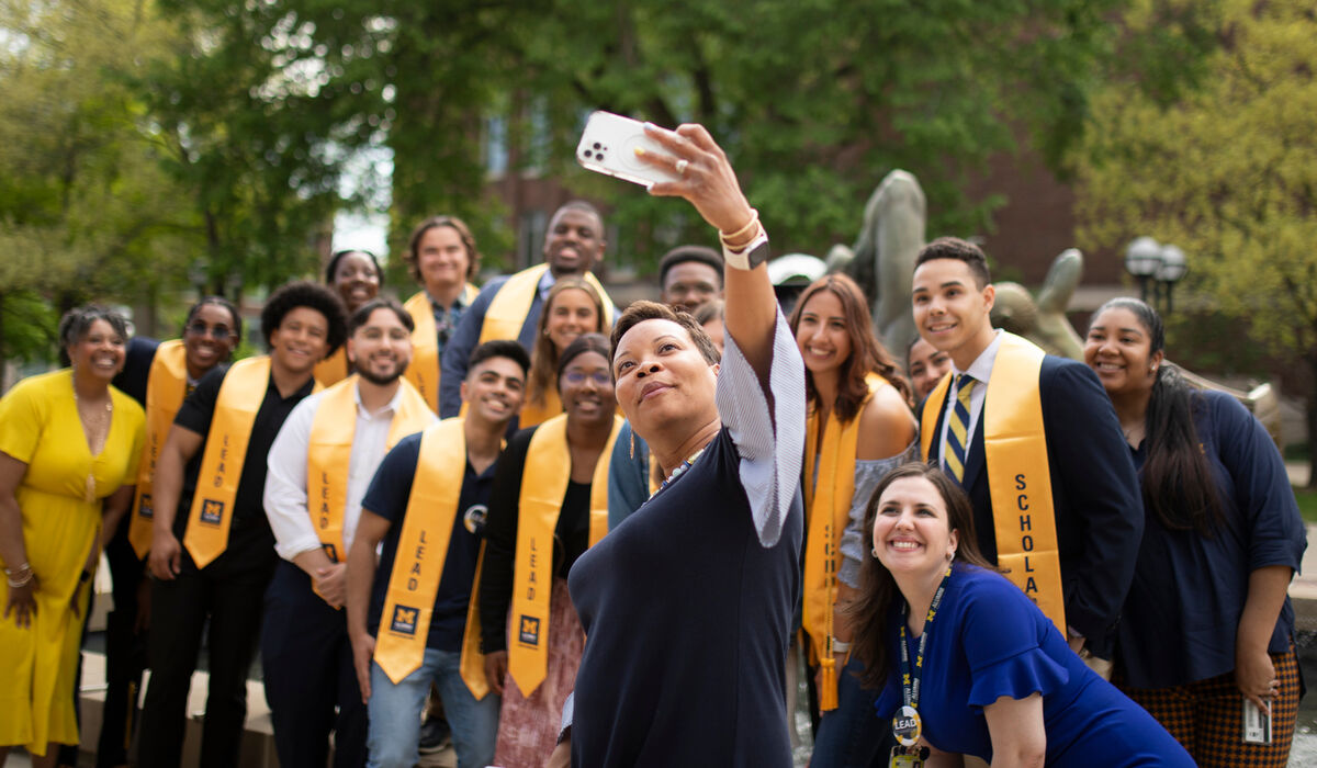 Woman holding a cell phone up for a selfie-style photo with a large group of college students and Alumni Association staff posed behind to fit in frame with her