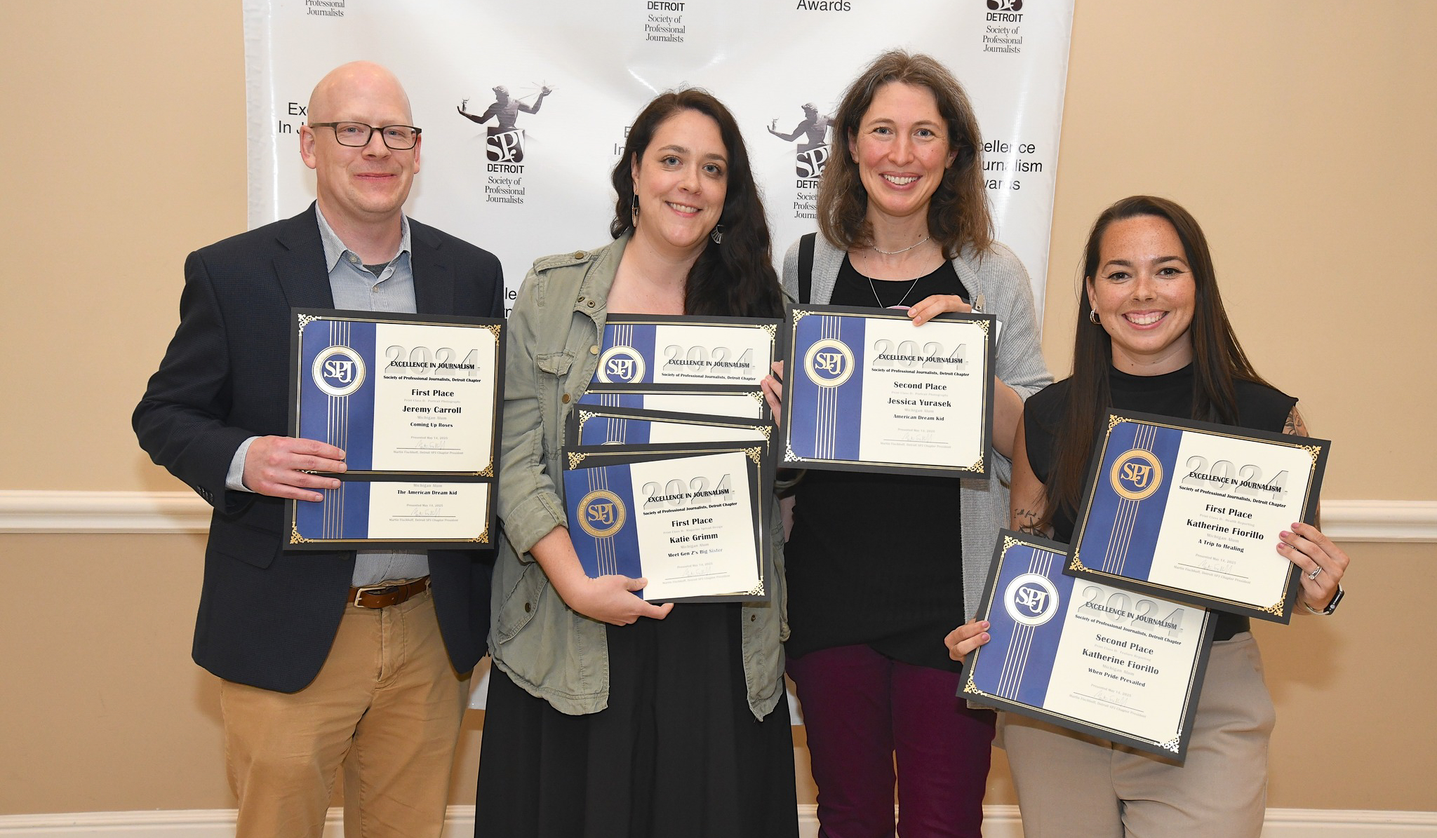 Four people hold various numbers of certificates in front of a step and repeat with the SPJ logo on it.