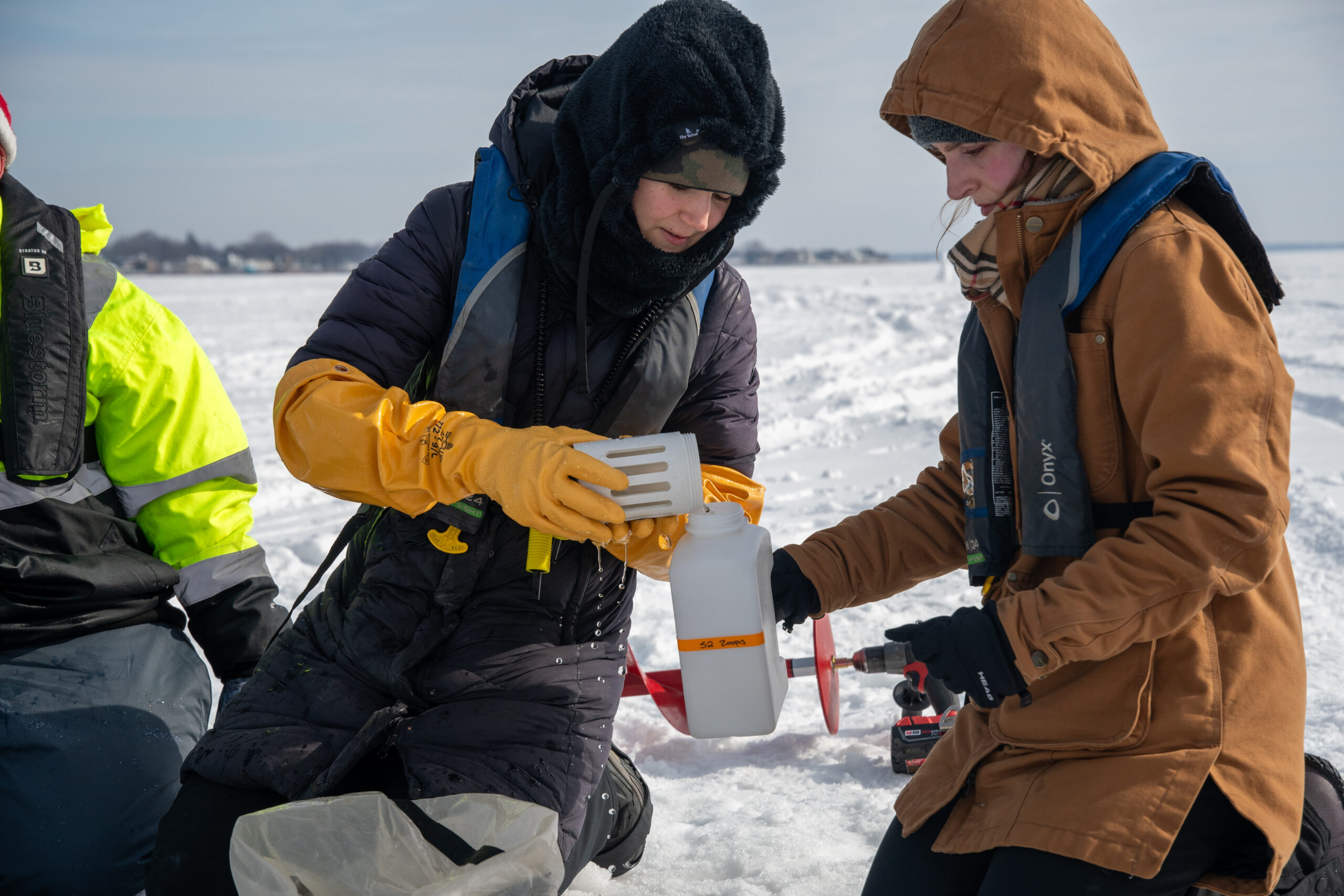 A person wearing large yellow gloves pours from one sampling container to another, held by a person in a brown winter jacket.