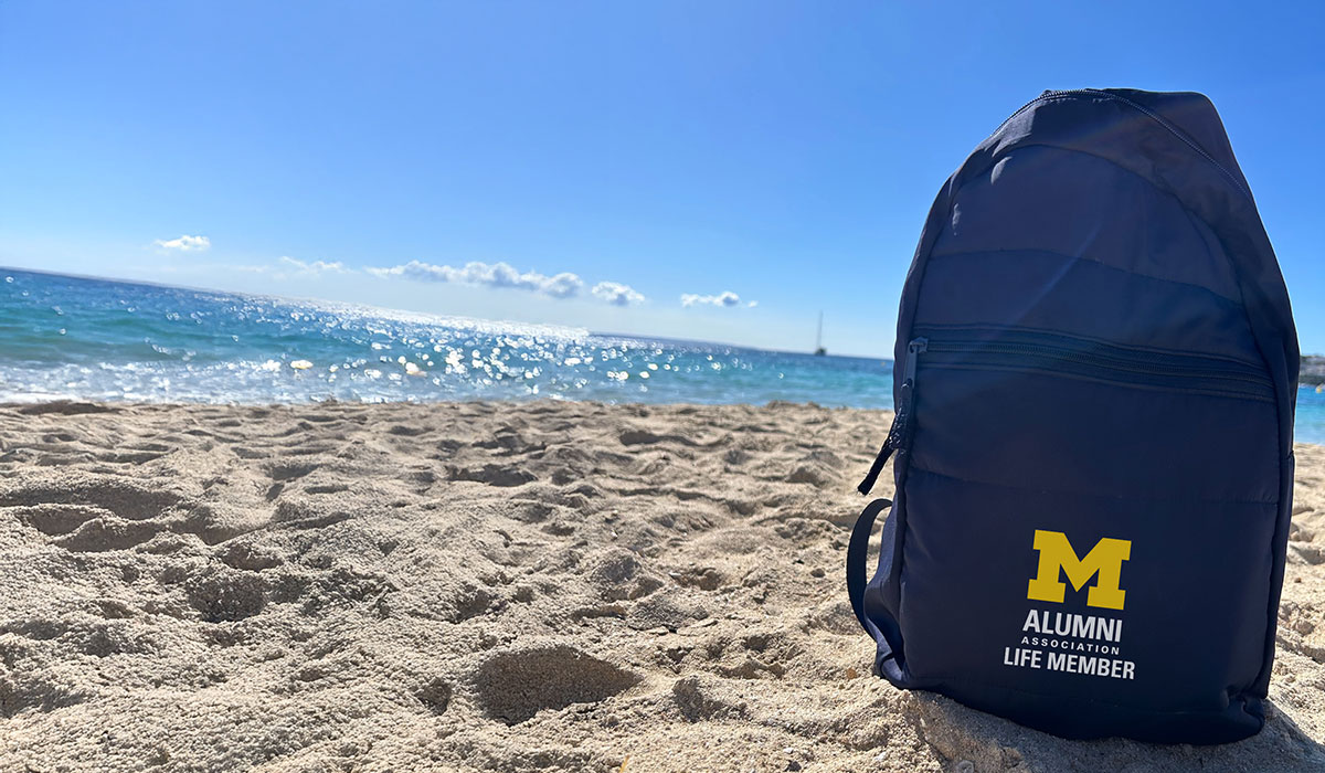 An Alumni Association Life Member backpack sits on a beach in front of a body of water.