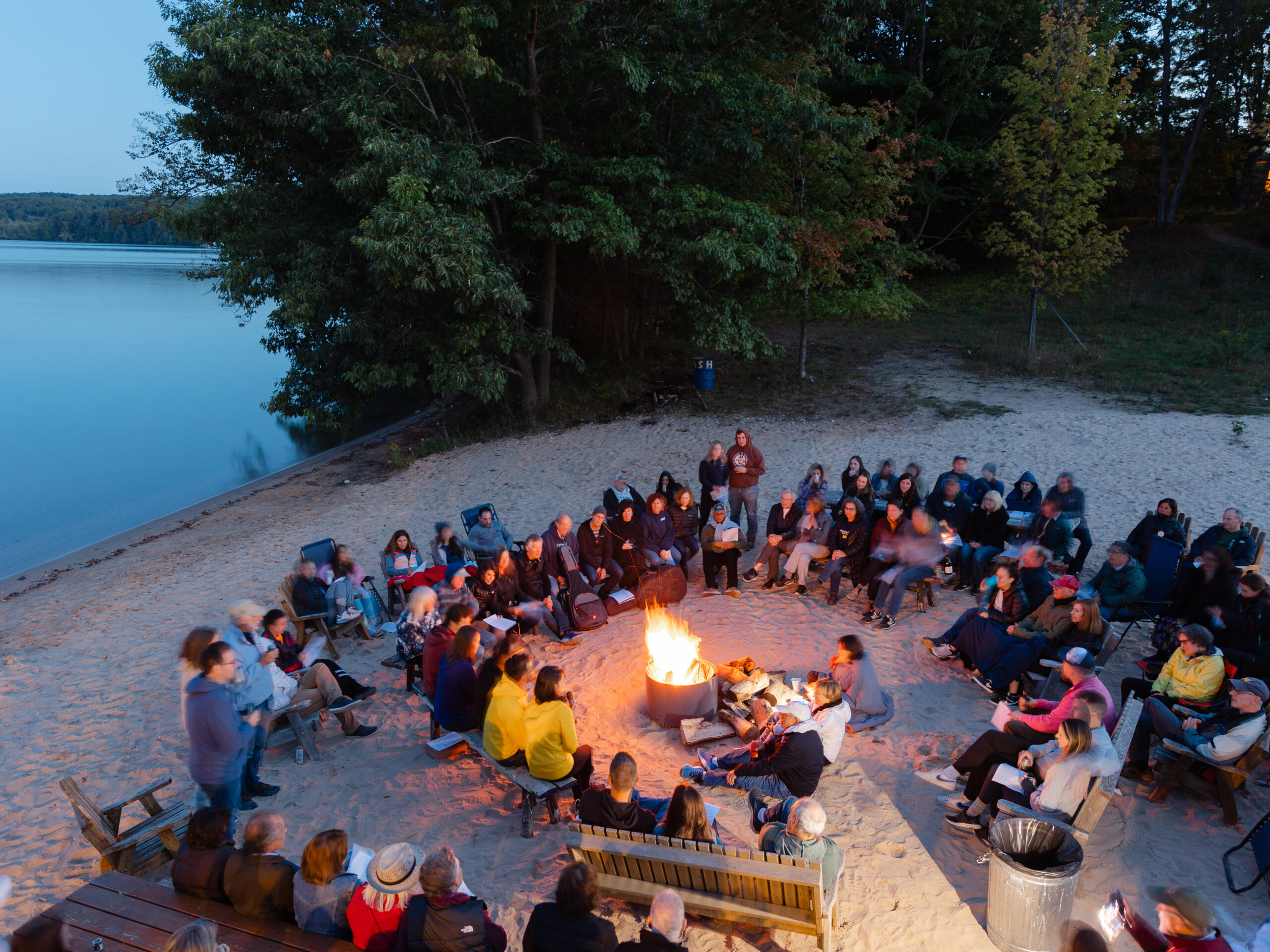 A large group gathers around a campfire on the beach of Walloon Lake at Camp Michigania. 