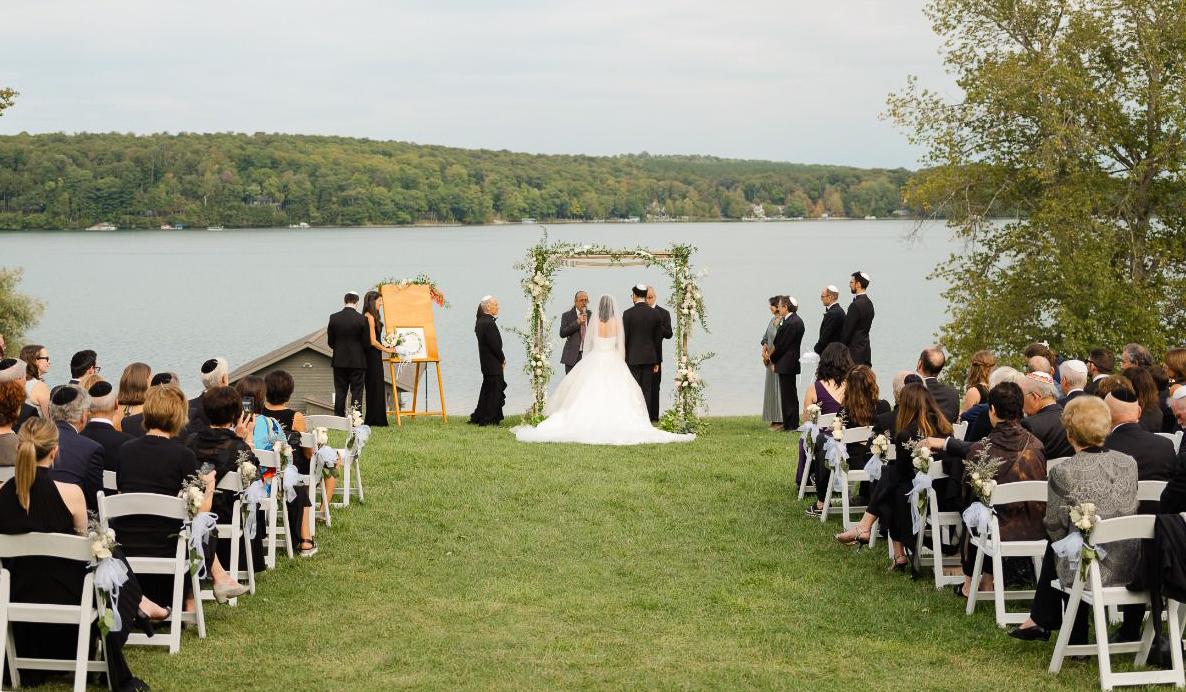 A bride and groom stand under a flowered archway overlooking Walloon Lake and green trees in the background. Seated are wedding guests.