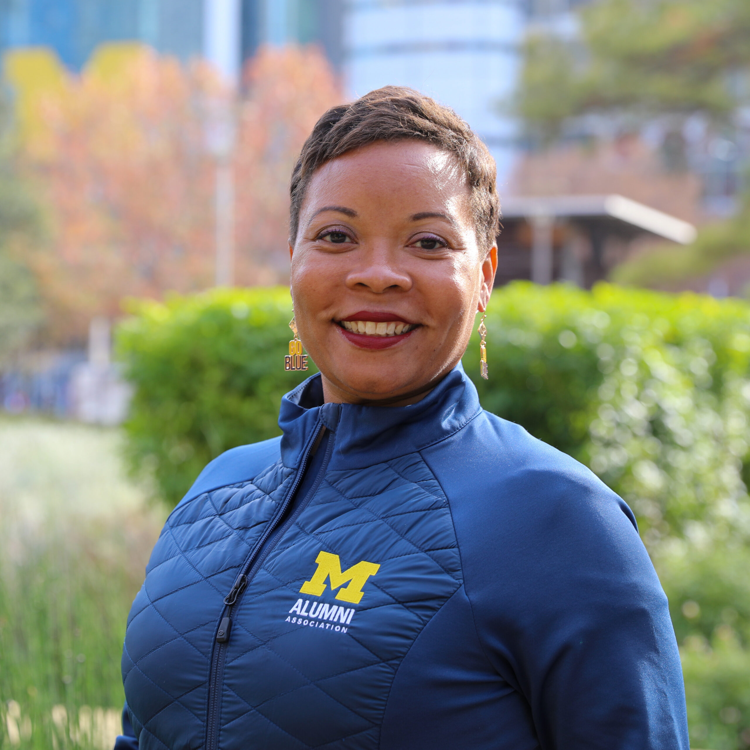 Headshot of McConnell, wearing an Alumni Association jacket and "Go Blue" earrings. The background has city buildings and greenery.