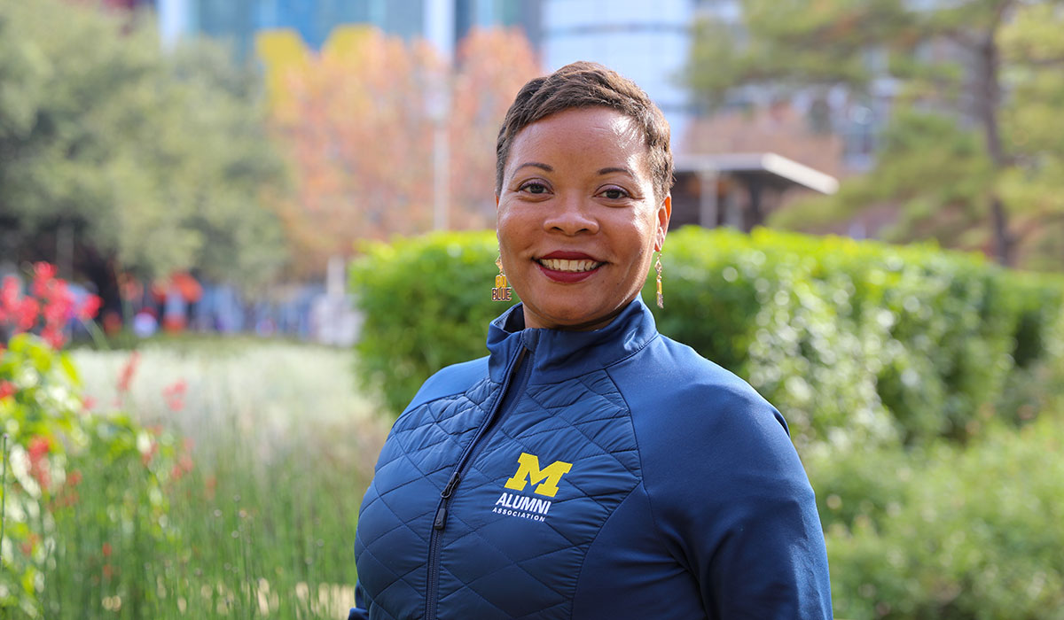 Headshot of McConnell, wearing an Alumni Association jacket and "Go Blue" earrings. The background has city buildings and greenery.