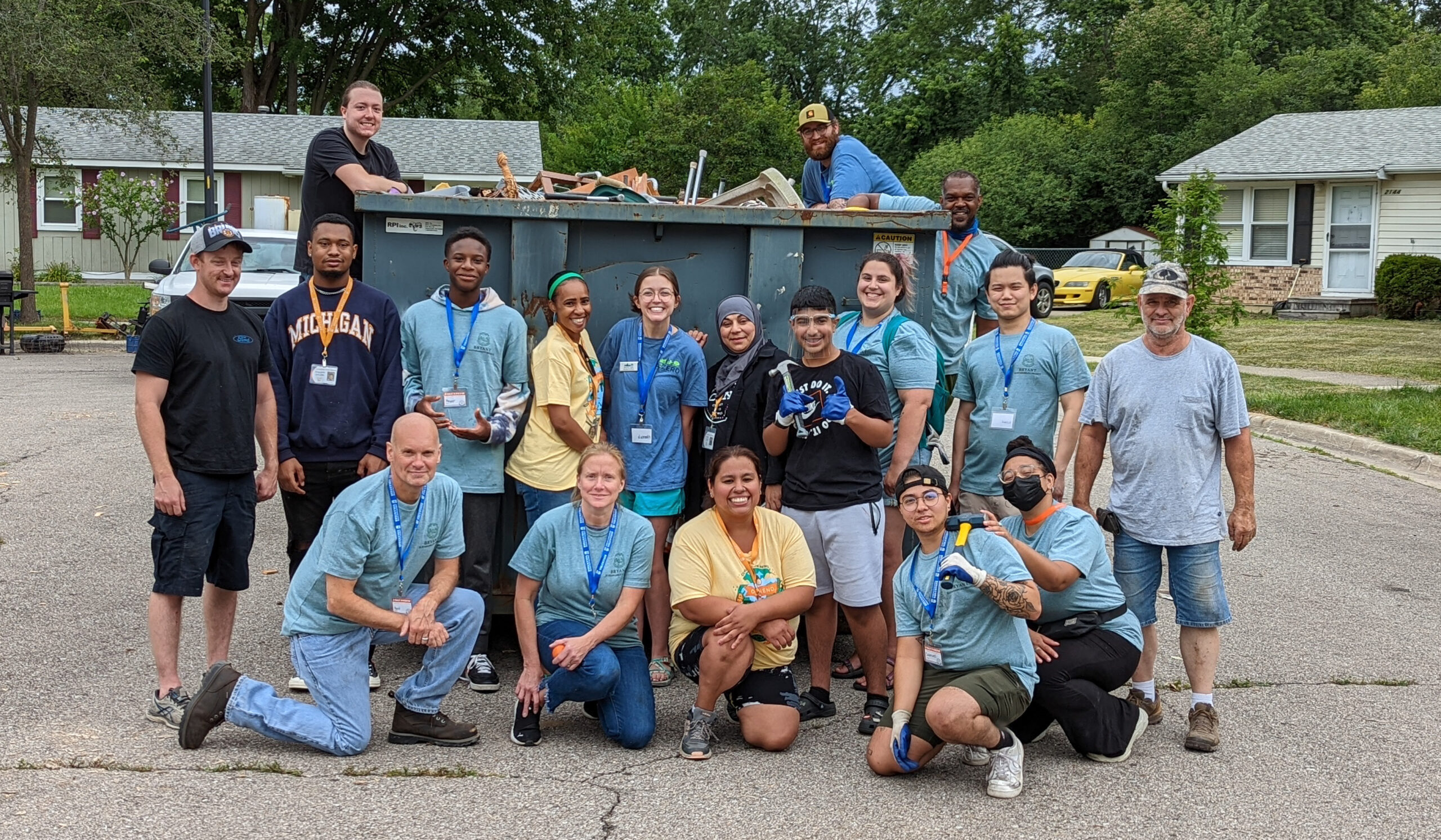 A large group of people in matching shirts stand in front of a dumpster on a neighborhood street.