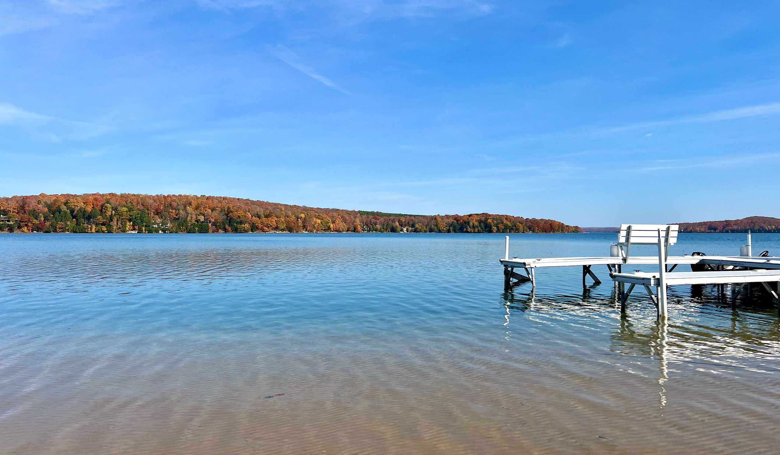 A quiet lake bordered by trees in full fall colors, with a white dock extending into the water under a clear blue sky.
