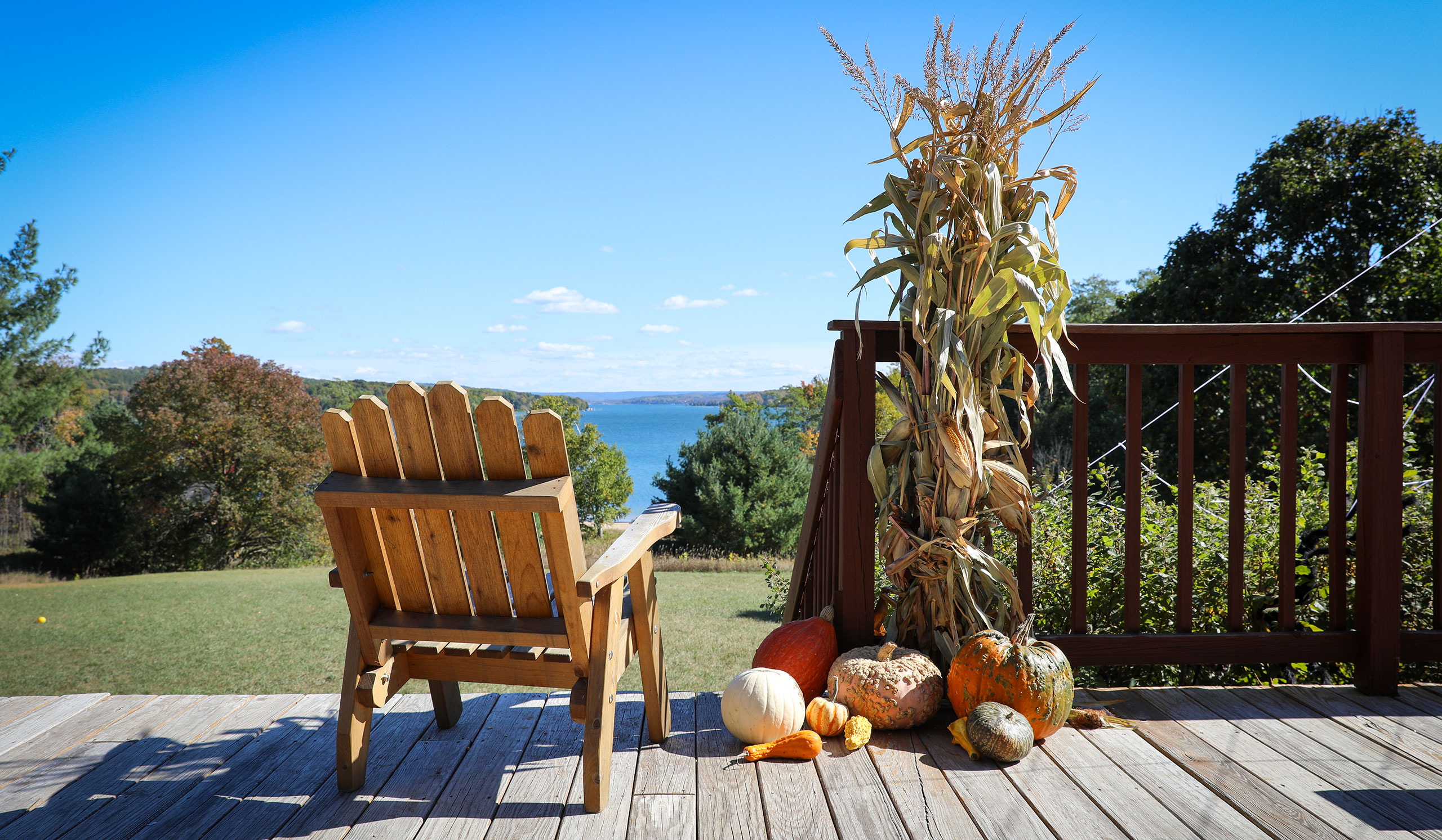 A wooden Adirondack chair sits on a deck facing a scenic lake view. Corn stalks and pumpkins are arranged beside the railing as autumn decorations.