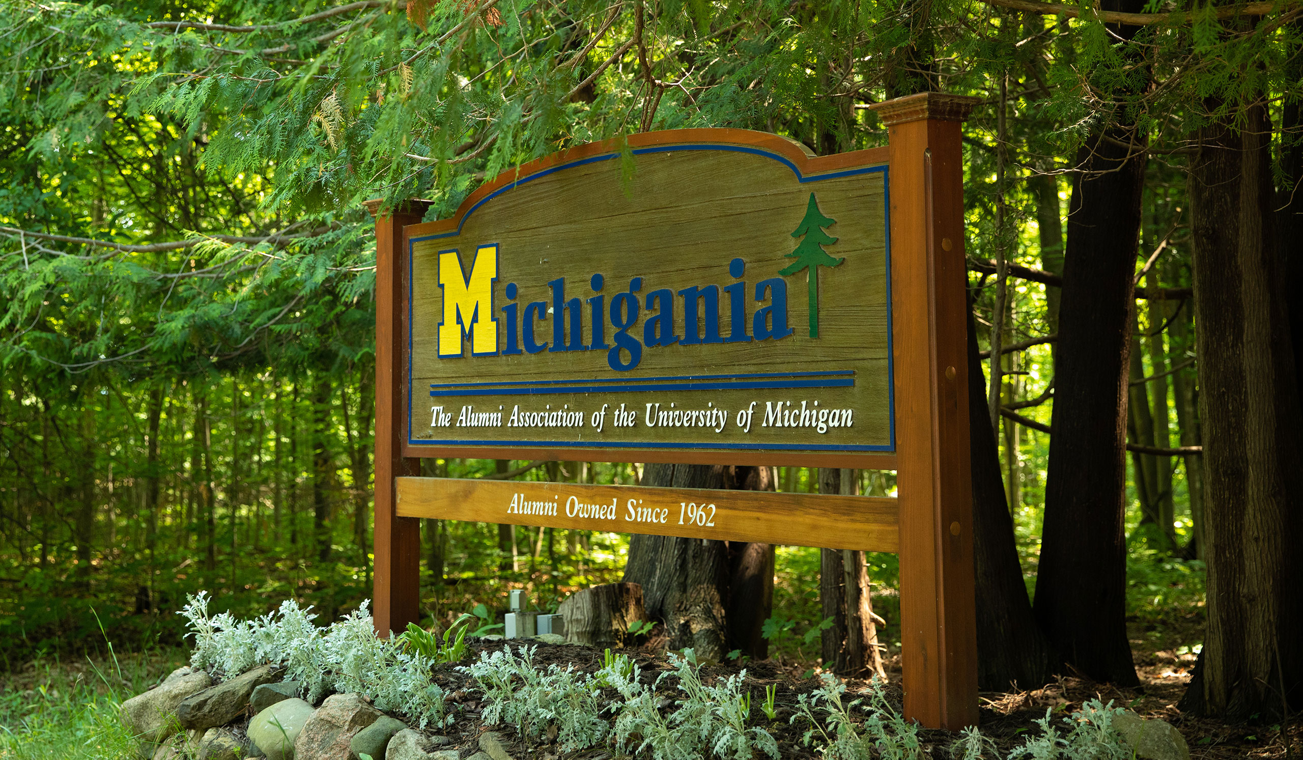 A wooden sign reads "Michigania" with a University of Michigan Block M and a green pine tree symbol, located in a wooded area. Below, it says the camp is alumni-owned since 1962.