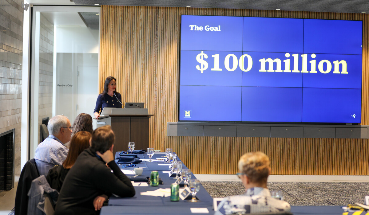 A woman stands at a podium next to a large blue screen which has the words, "The goal: $100 million" on it. There are a number of people sitting at a table listening to the speaker.