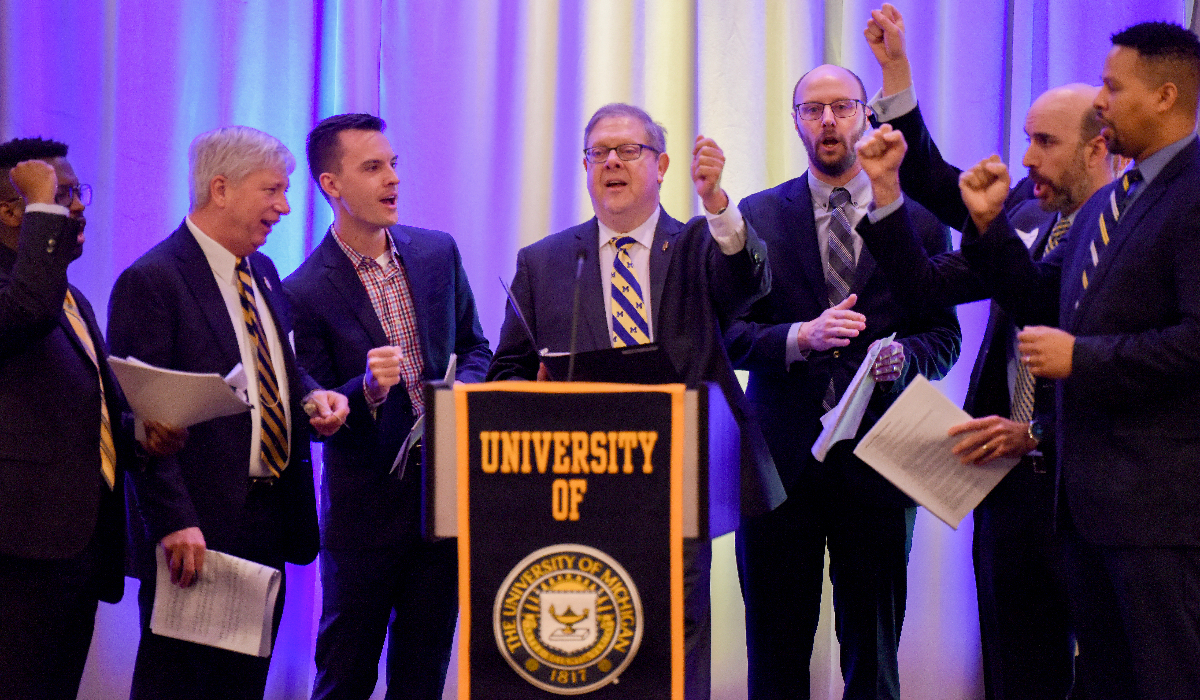 U-M alumni sing Hail to the Victors on a stage with a podium that says "University of Michigan."