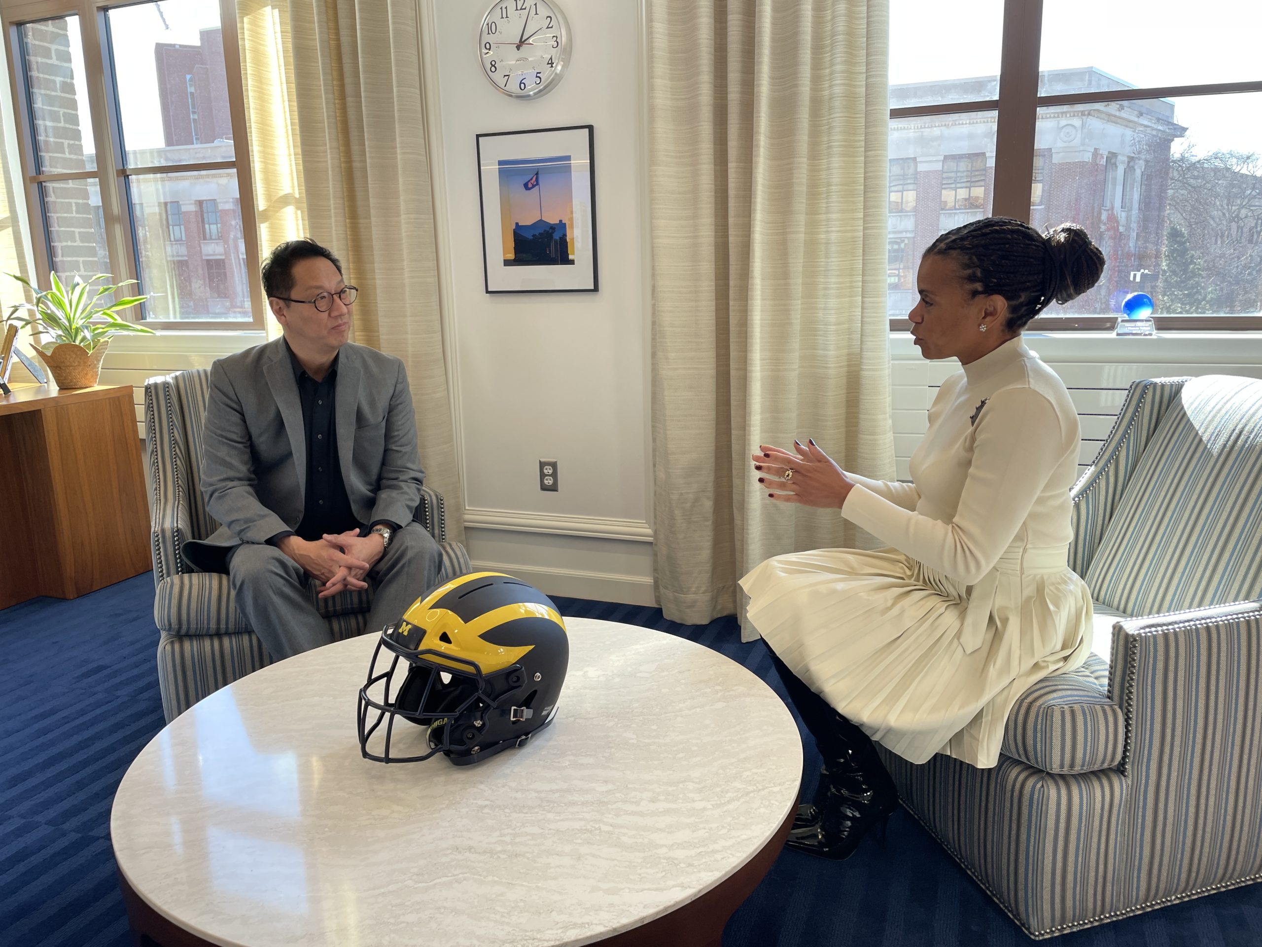 Corie Pauling sits in an open office space with U-M President Santa Ono. Ono is wearing a grey suit and Corie is wearing a white, long-sleeve dress. They are sitting in arm chairs with a round table between them.