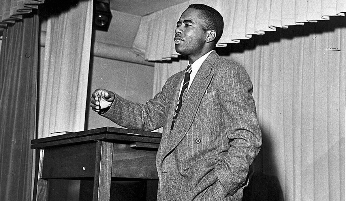 Black and white photograph of Paul B. Cornely lecturing to an unseen audience from a stage with curtains in the background. Cornely is an African American man with close-cut hair, dressed in a suit with pinstripes