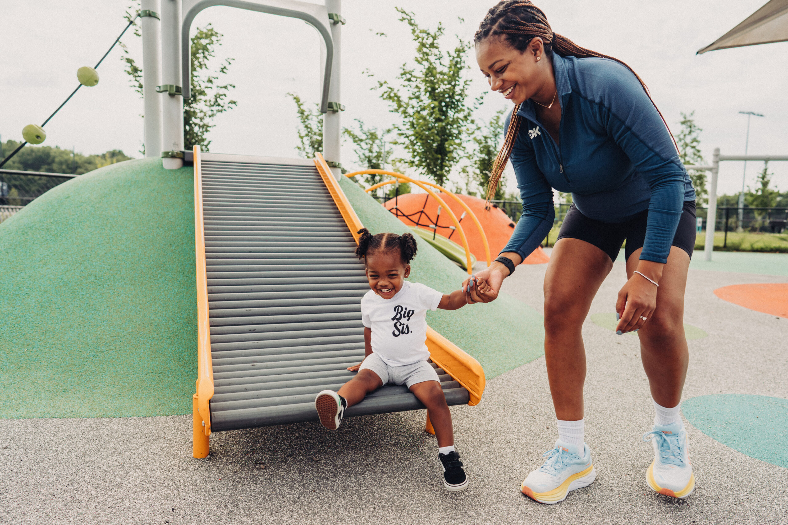 Ashlee Greene helps her daughter down a slide in a park. Ashlee is wearing a blue/grey athletic top, black shorts, and white shoes. Her daughter is dressed in a white outfit.