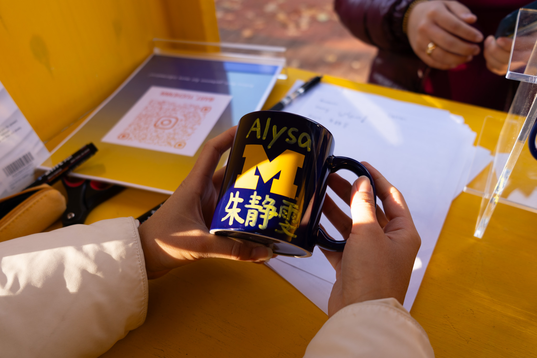 Yuchen Wu holds a maize and blue U-M mug with "Alysa" written in English and the student's mother language. 