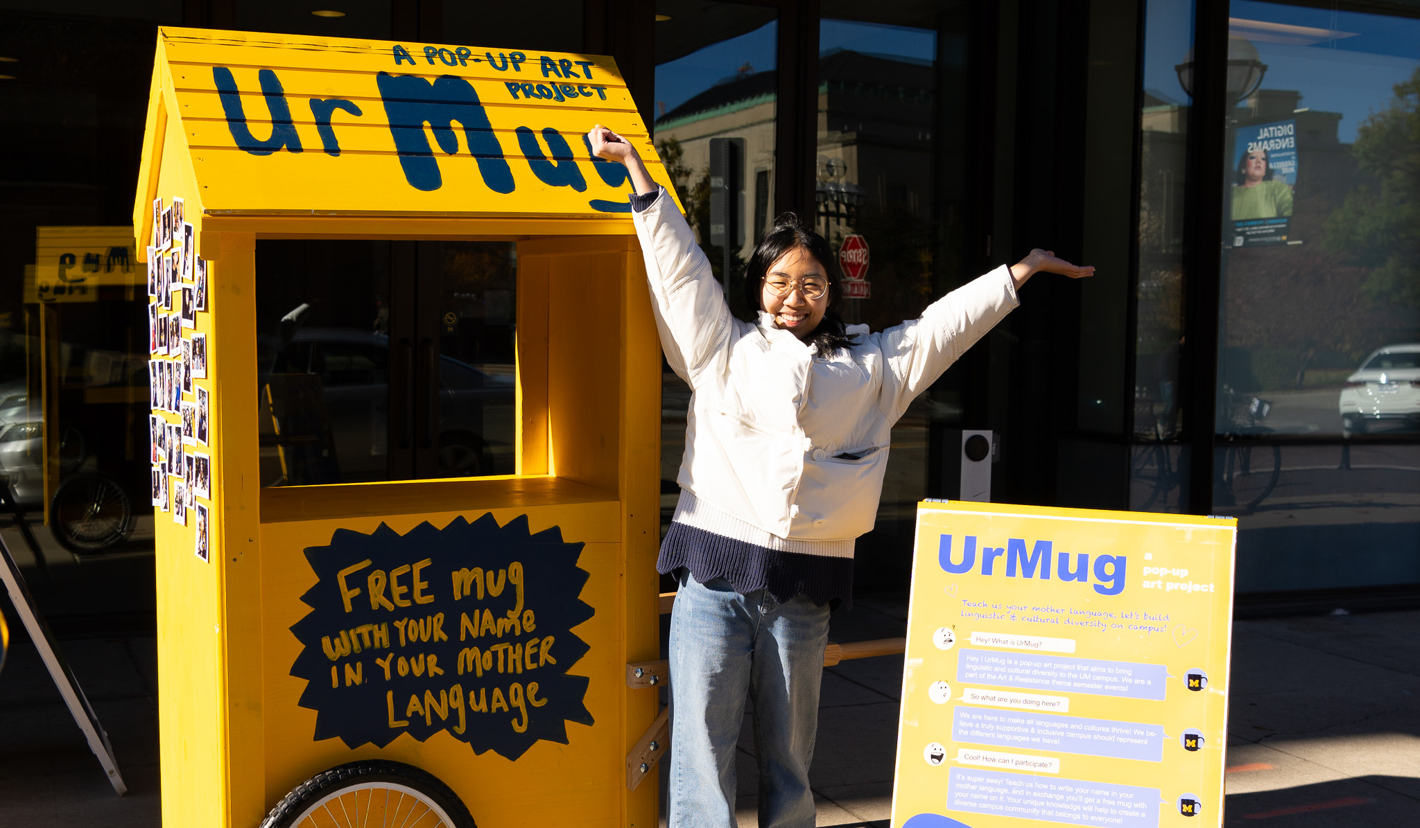 Yuchen Wu, wearing jeans and a white jacket, poses with her yellow "Ur Mug" cart and a sign describing her installation.