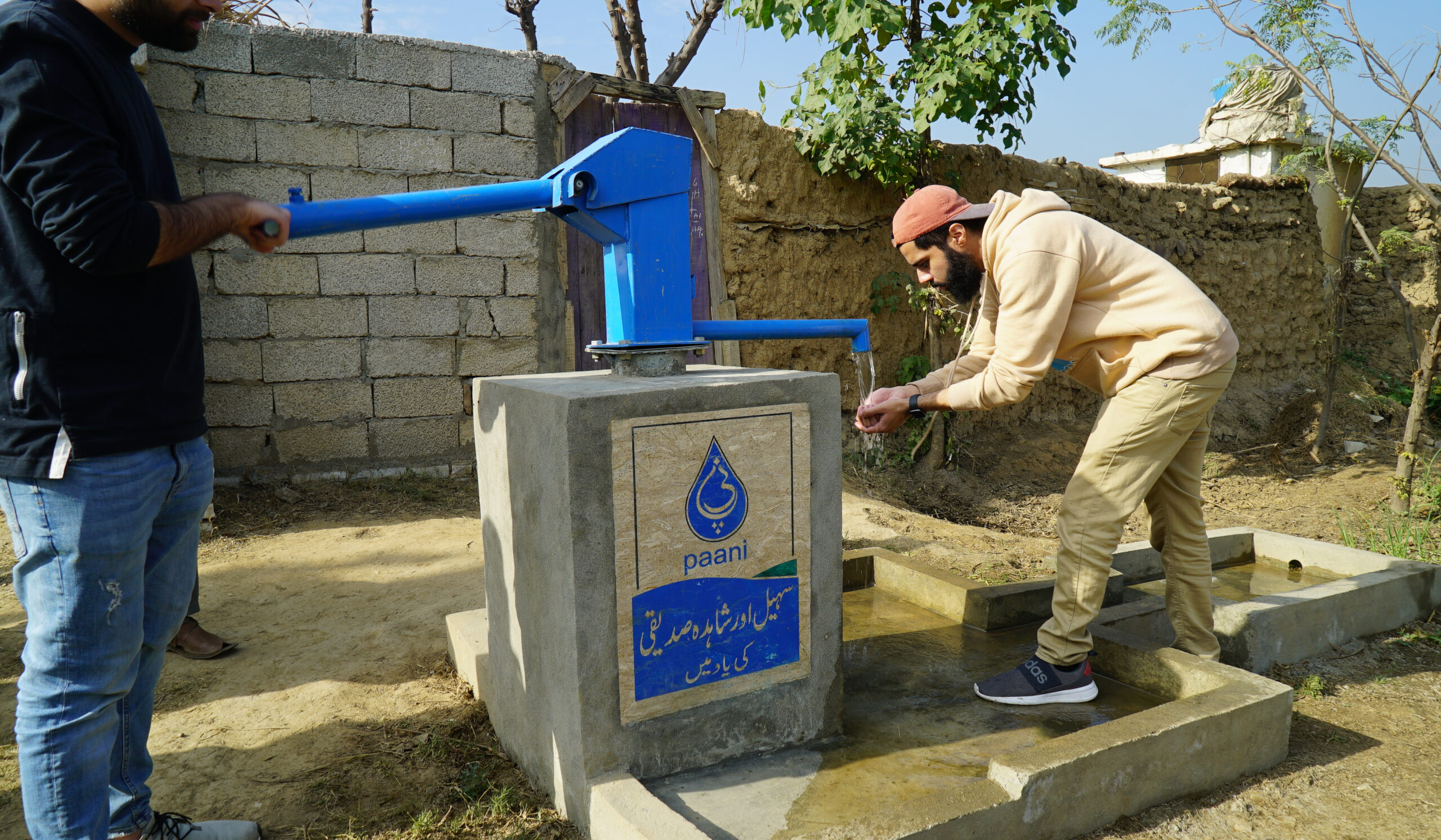 A person in jeans and a dark shirt pumps a Paani water well while a person in tan pants and a pan hoodie cups his hands at the other end of the well.
