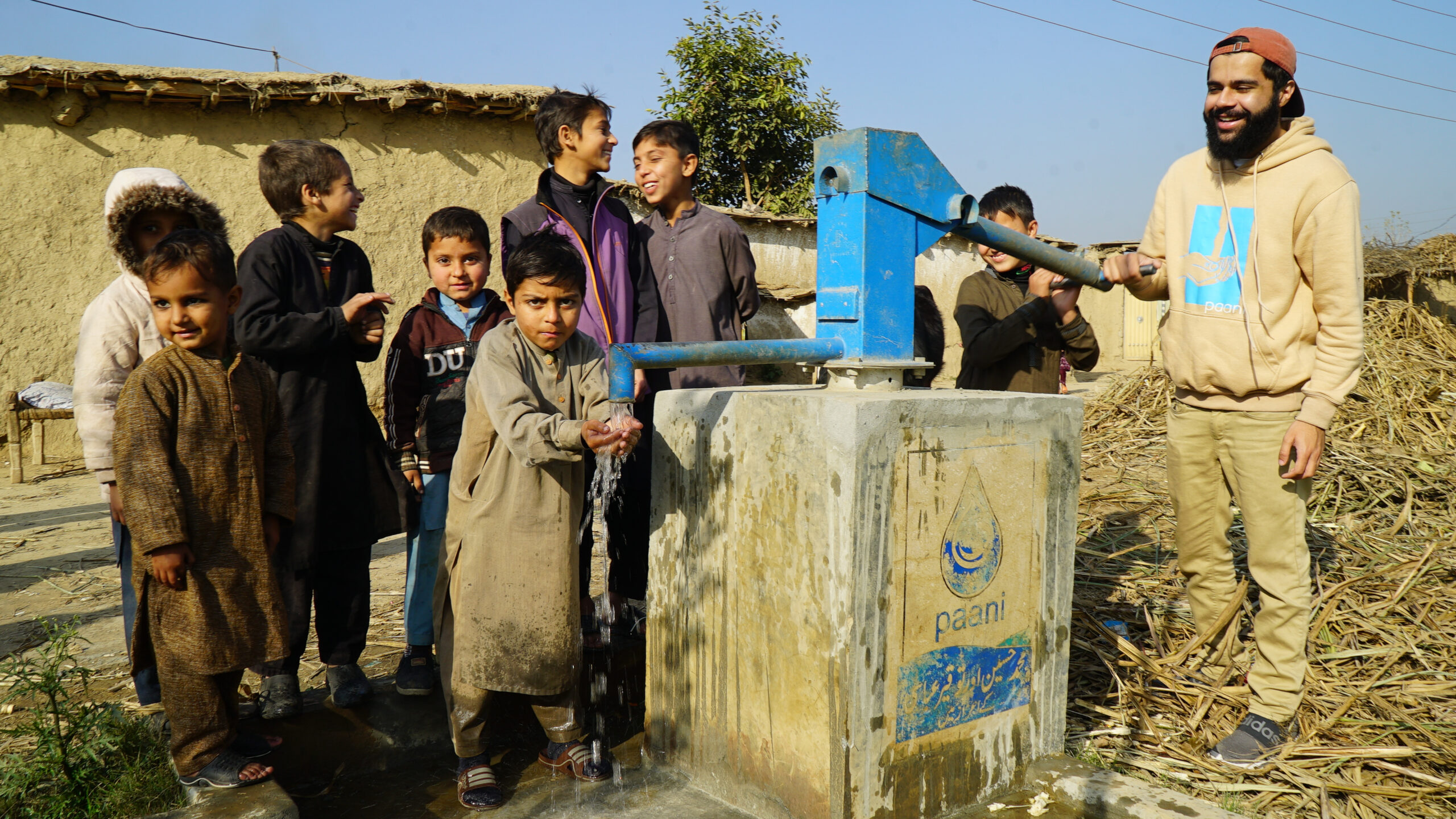 A group of children stand around a Paani well while an adult wearing a tan sweatshirt and pants pumps the water from the well. 