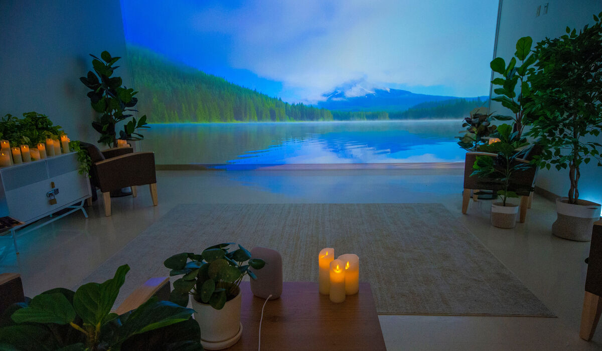 Interior of a Recharge Room, overly dark with plants and an artificial candle in the foreground and the main background being a projection of a serene mountain lake and forest scene.