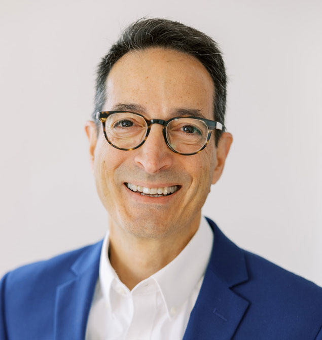 A headshot of Damian Zikakis, a man in glasses, a white dress shirt, and a blue jacket.