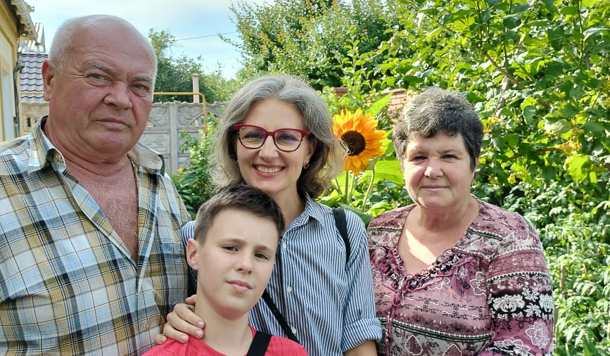 A young boy stands in front of three people. From left to right: an older bald man with a serious expression in a plaid shirt; a smiling woman with salt-and-pepper hair, red glasses, and a striped shirt; and a woman with short salt-and-pepper hair and a pink and purple shirt. The background is mostly green with a sunflower.