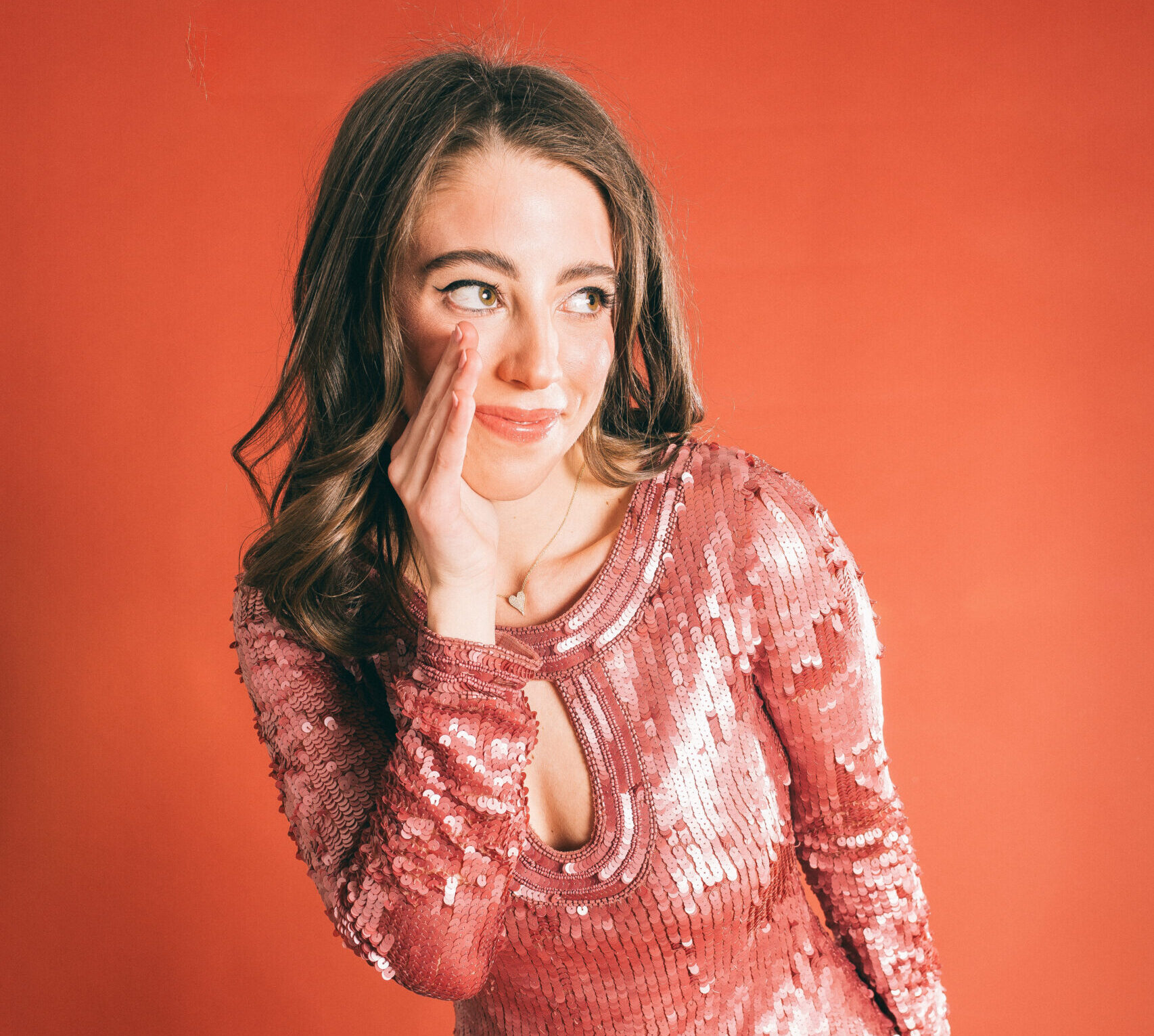 Eli Rallo, in a sparkly pink dress, stands in front of a red background with her hand cupped to the side of her mouth as if telling a secret.