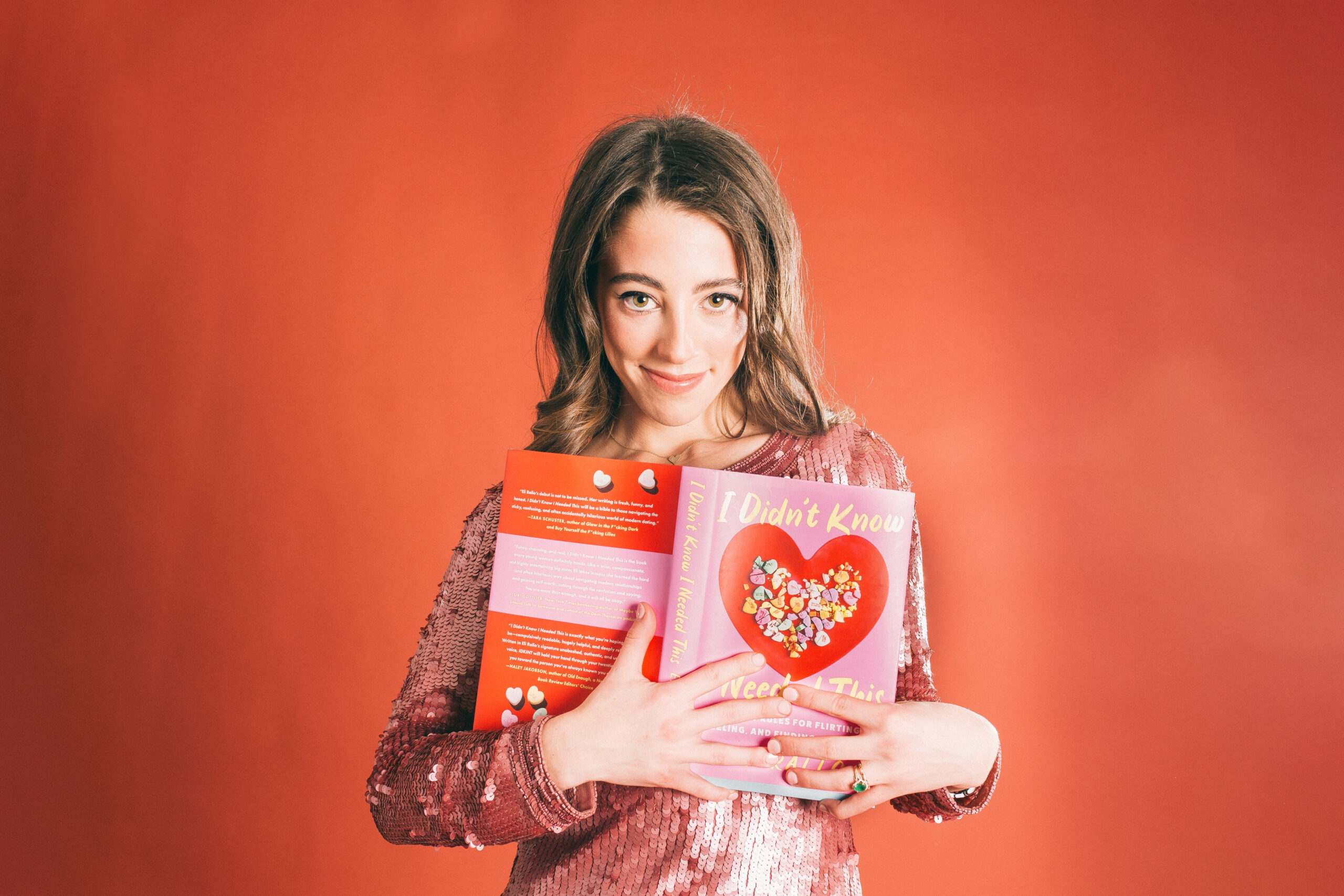 Eli Rallo in a sparkly pink dress, holds her open book to her chest in front of a red backdrop.