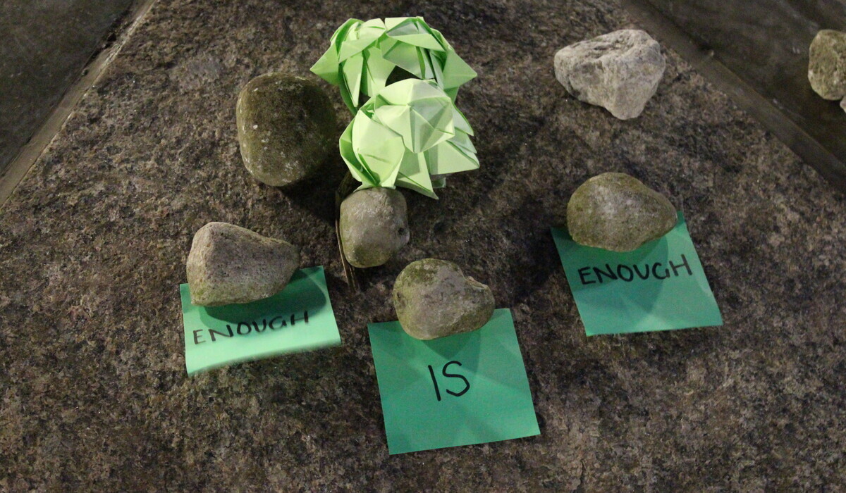 Three green pieces of paper reading "Enough is enough" are placed under rocks. Two other crumpled pieces of green paper lay above them. 