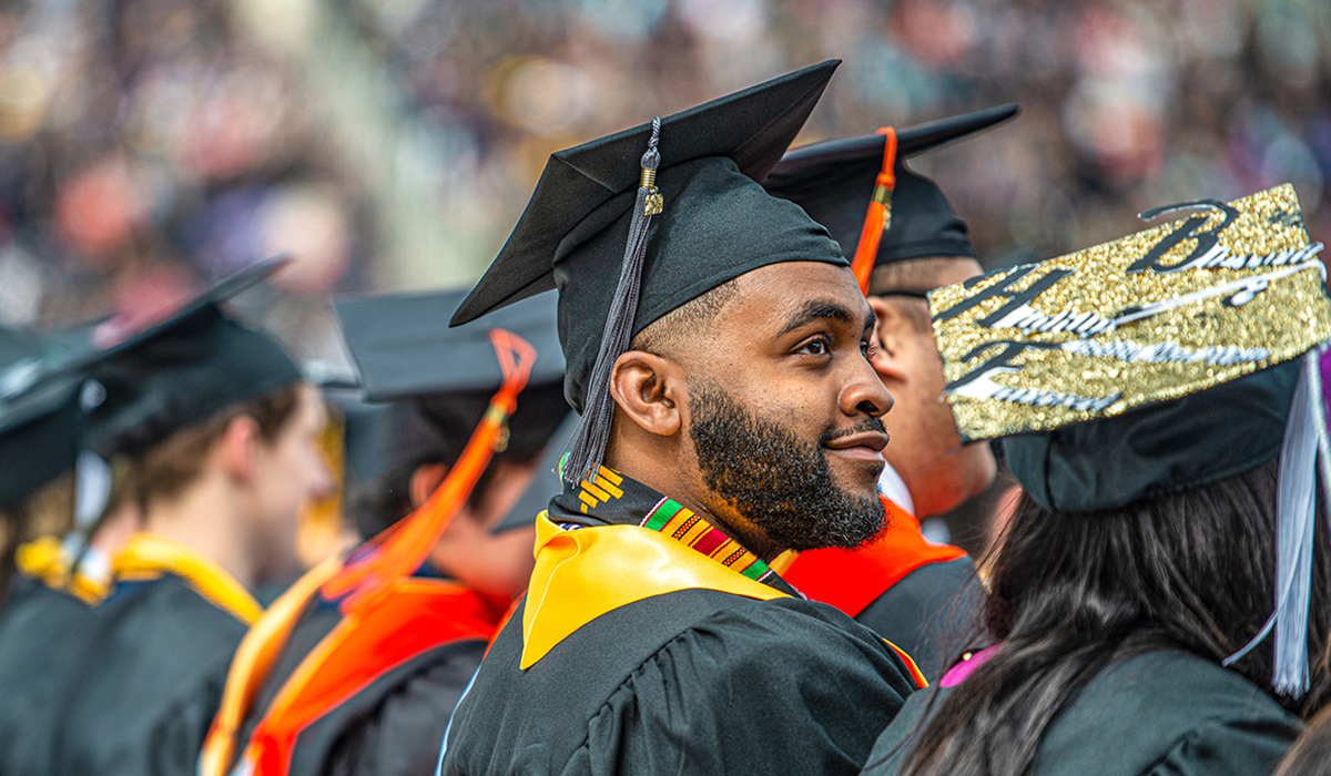 Graduates wearing caps and gowns at the University of Michigan's Spring Commencement ceremony. One graduate, seen from the side, looks forward with a proud expression.