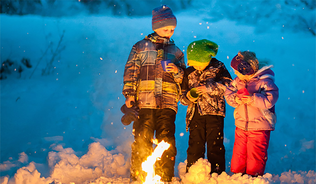 Three kids huddle around a campfire in the middle of a snowy landscape.