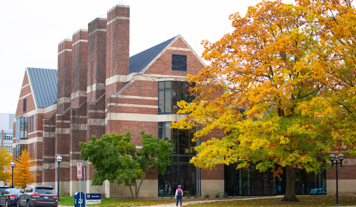 The Alumni Center is partially blocked by a large treat that has various colors in the fall. The tree has red, yellow, and green leaves.