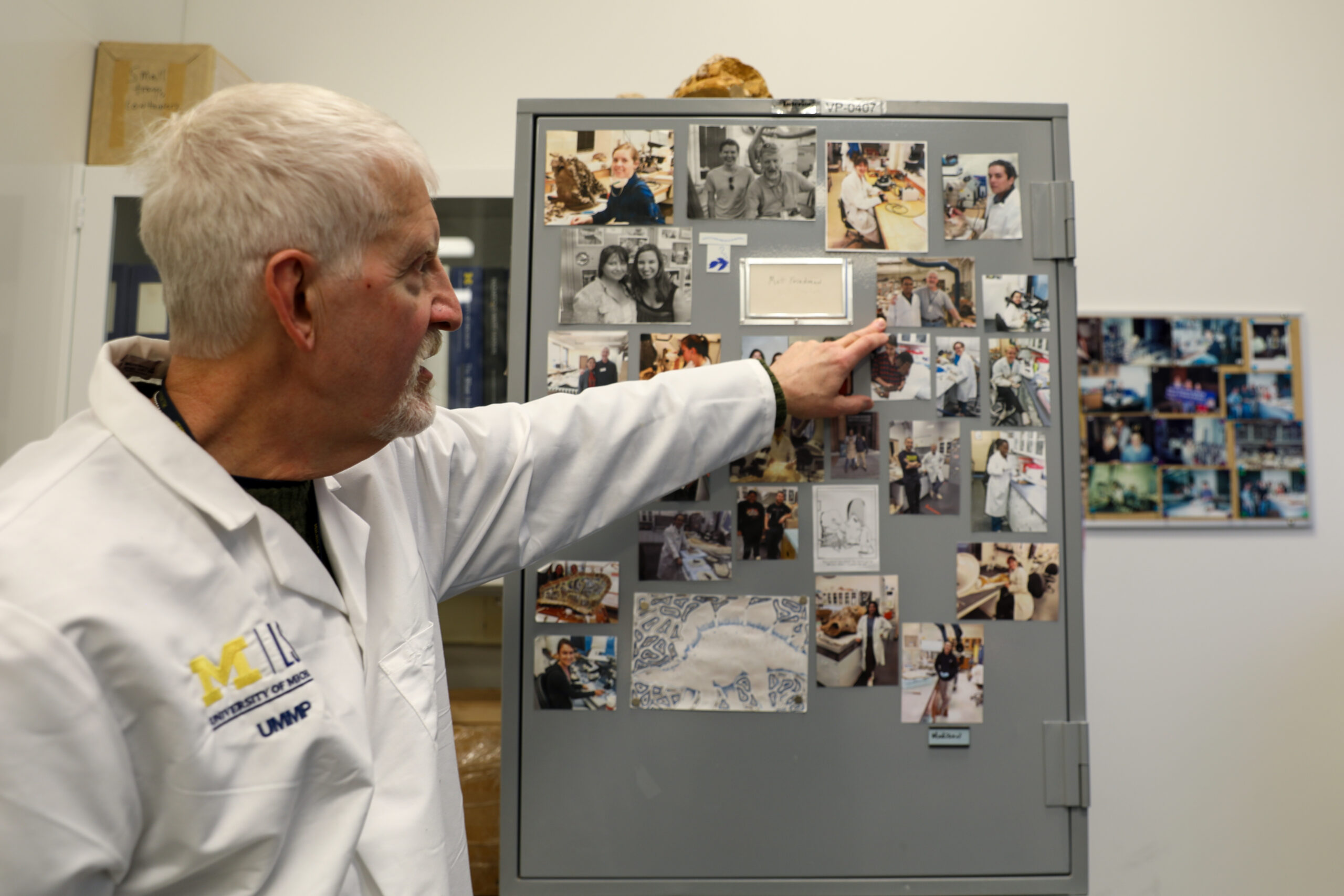 Chief Vertebrate Preparator Bill Sanders points to a photo among a collage of photos of former students who worked in the lab.