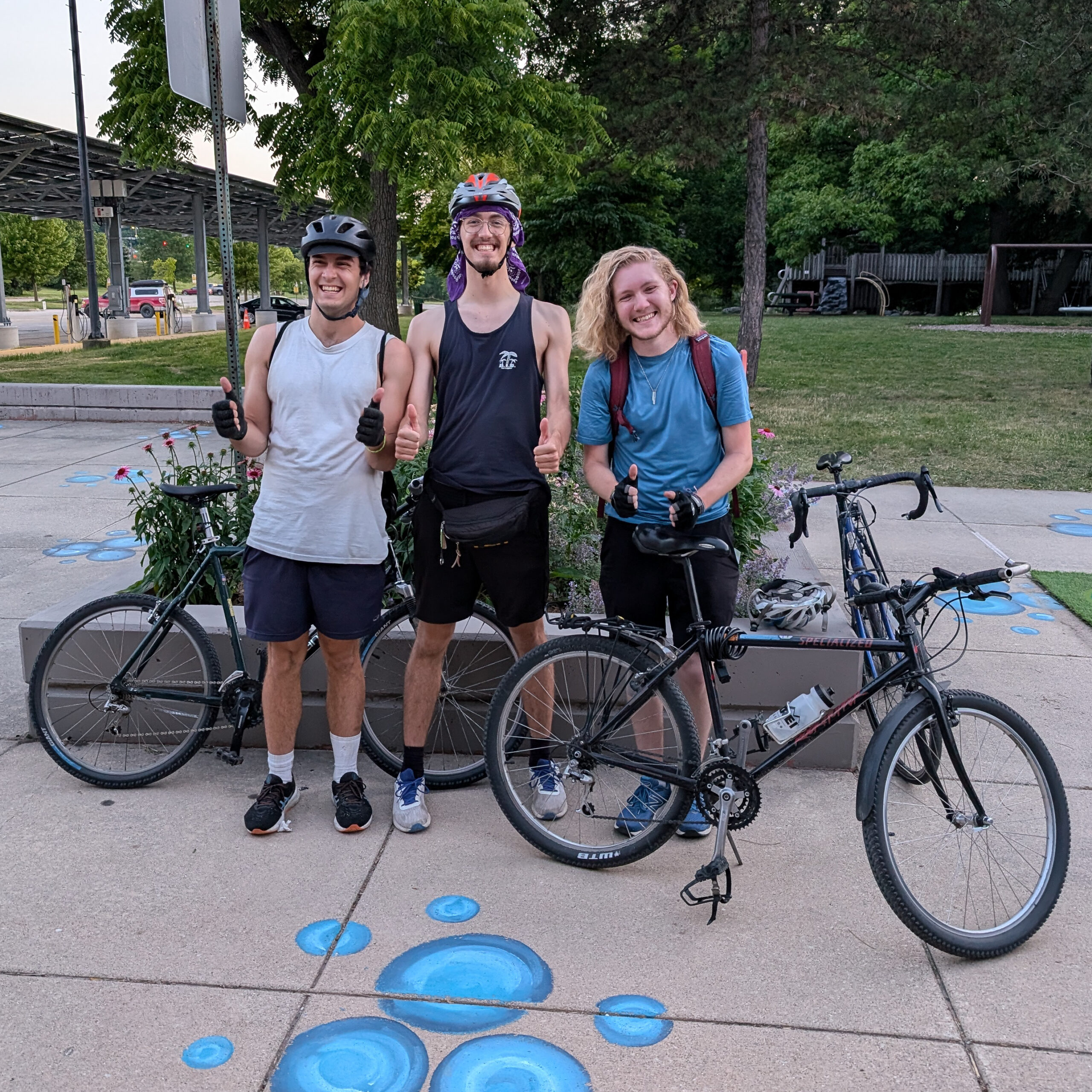 Three people pose on a sidewalk with chalk drawings on it. They are standing with three bikes, two are wearing helmets, and all are smiling and giving thumbs up.