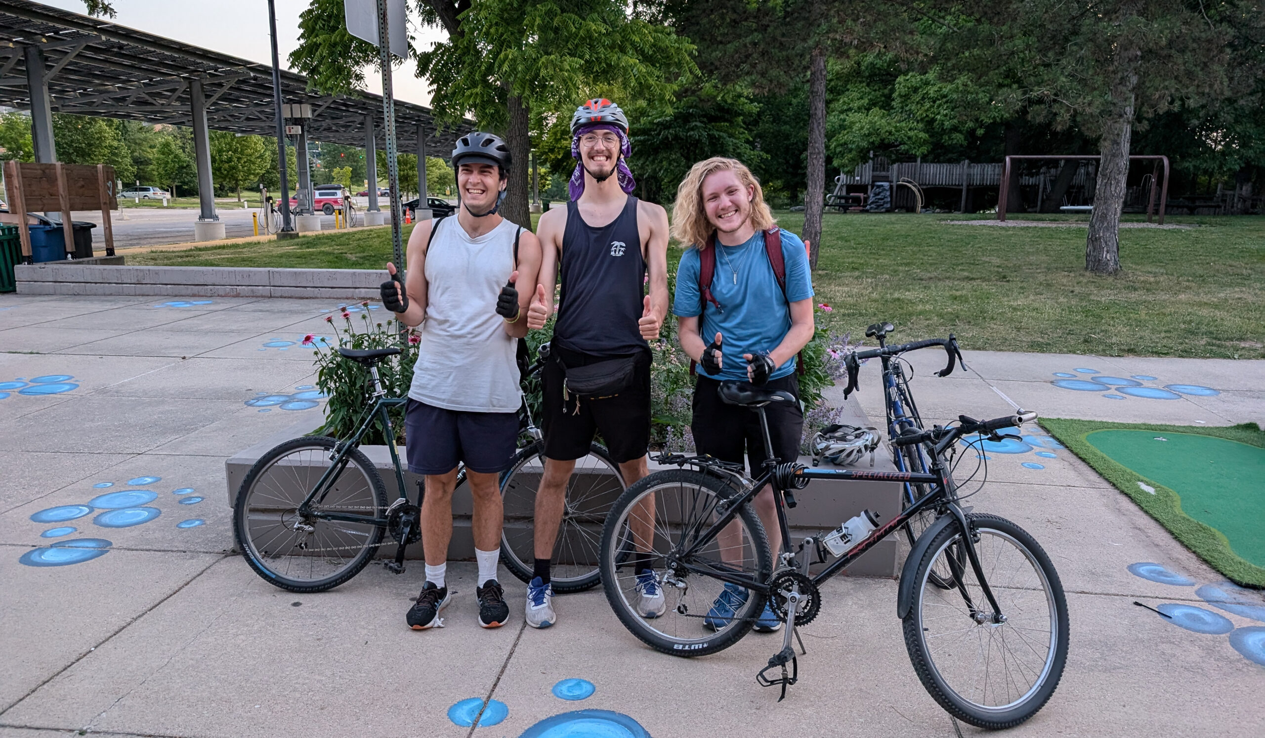 Three people pose on a sidewalk with chalk drawings on it. They are standing with three bikes, two are wearing helmets, and all are smiling and giving thumbs up.