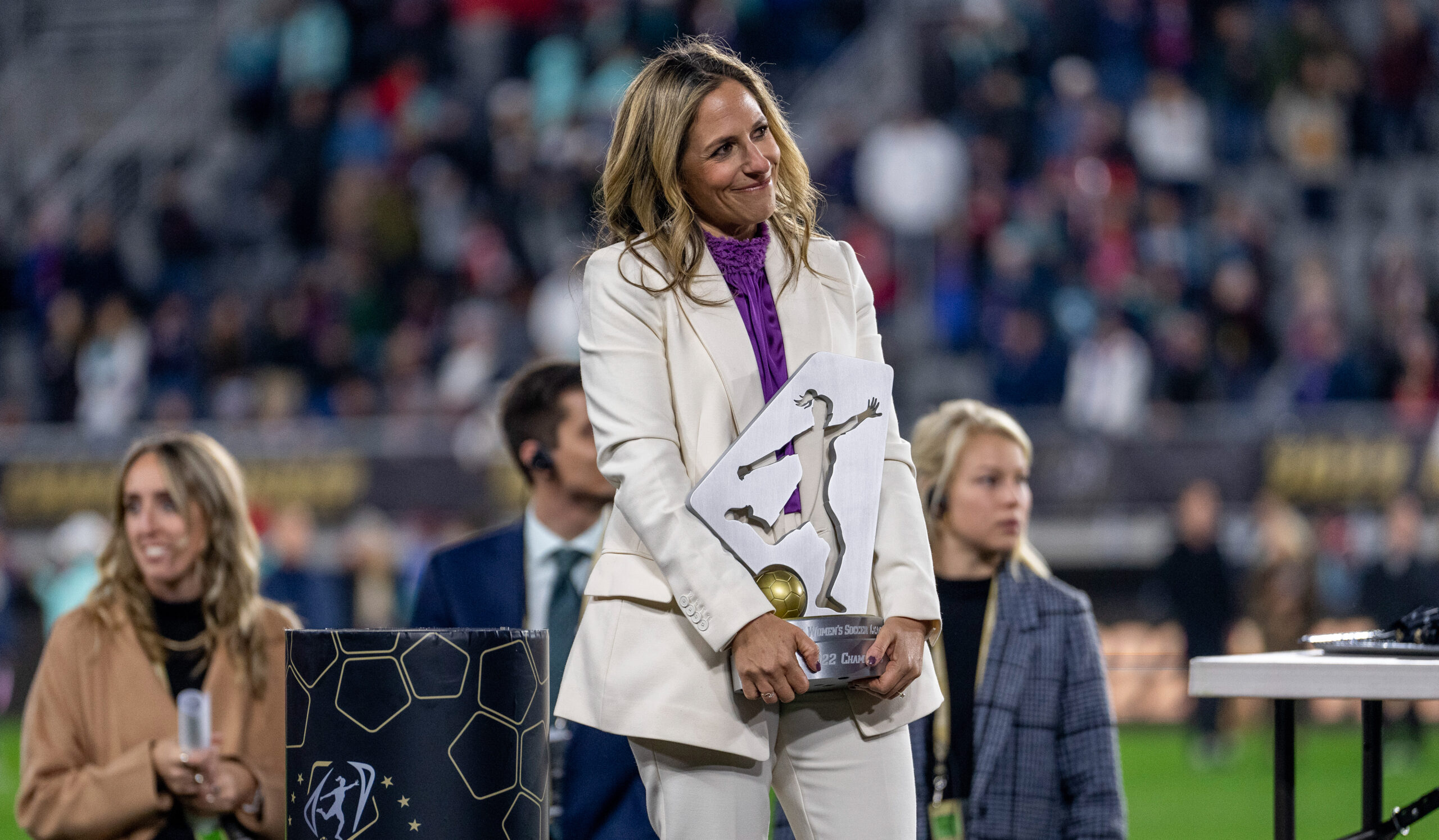 Jessica Berman, in a white pant suit and purple shirt, holds the NWSL championship trophy and is looking off to the right.