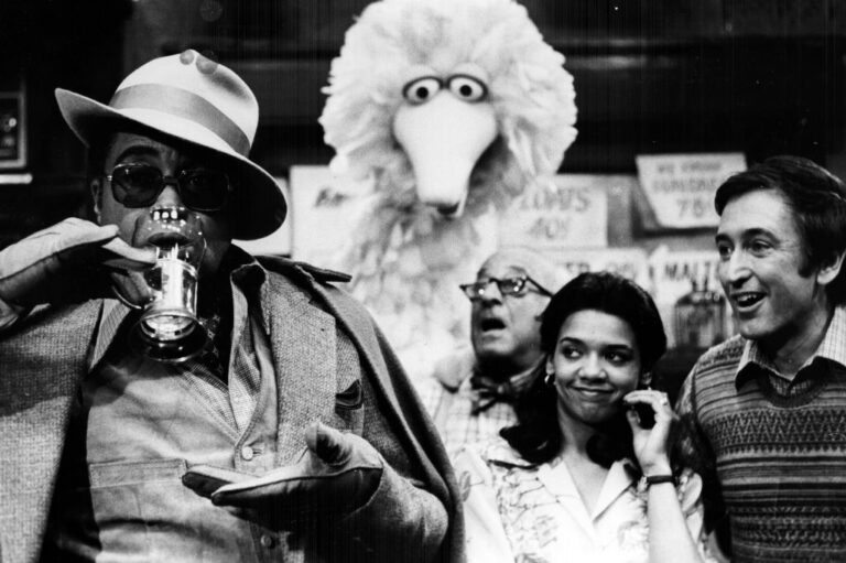 Black-and-white photo of James Earl Jones, dressed in expensive gentlemen's attire, delicately taste-testing an egg cream soda as the cast of Sesame Street look on in anticipation.