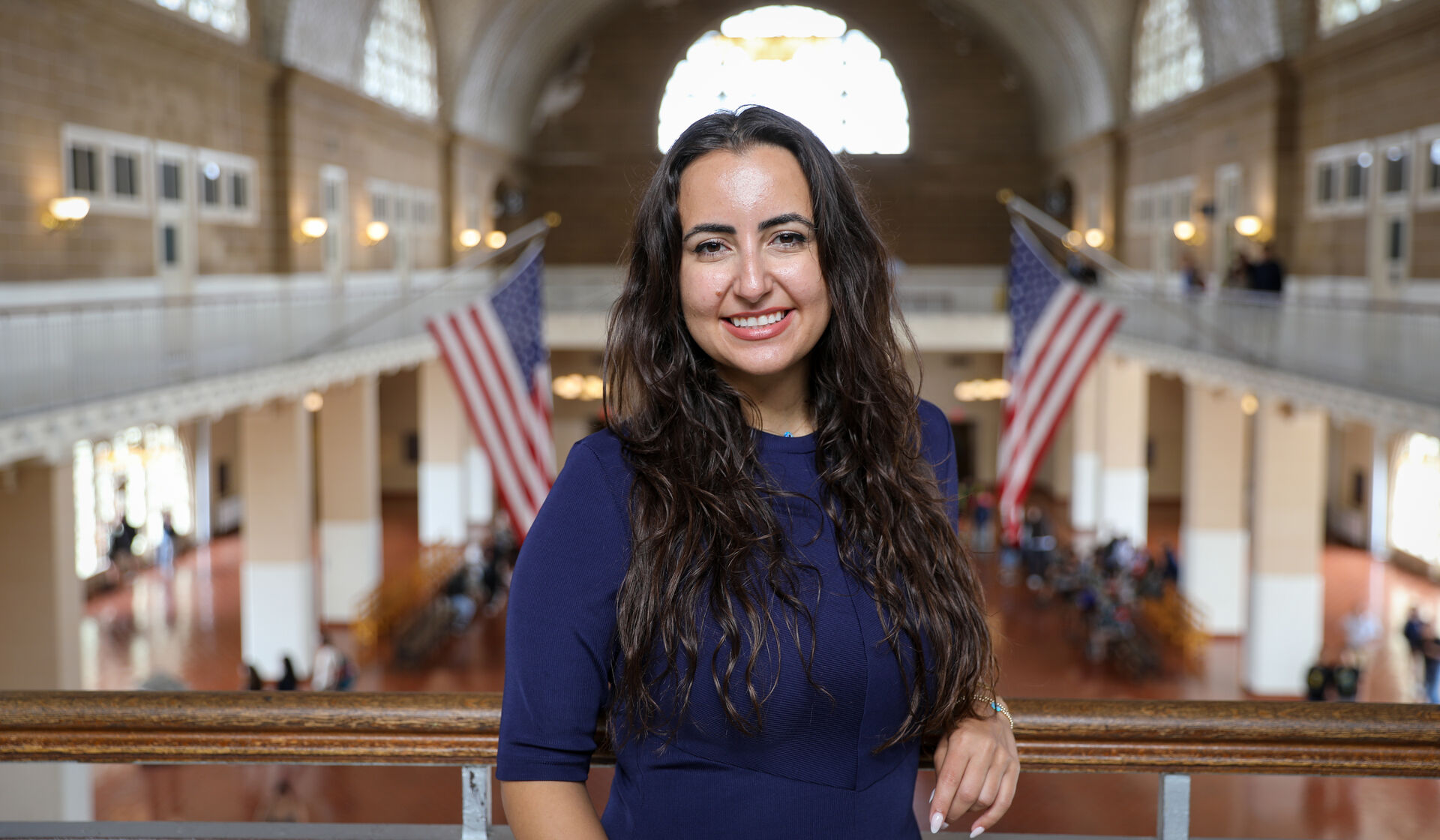 Ghida Dhager poses for a photo portrait within the Ellis Island National Museum of Immigration. Notably, two U.S. flags, positioned on the walls in the background, flank her sides.