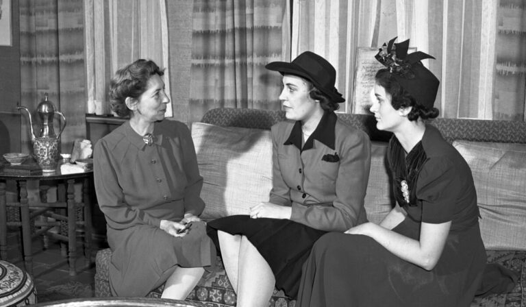 Black and white photo of three women sitting on a sofa in a home parlor-style room, conversing with each other in a slightly posed way for the camera, dressed in formal1930's-style attire.