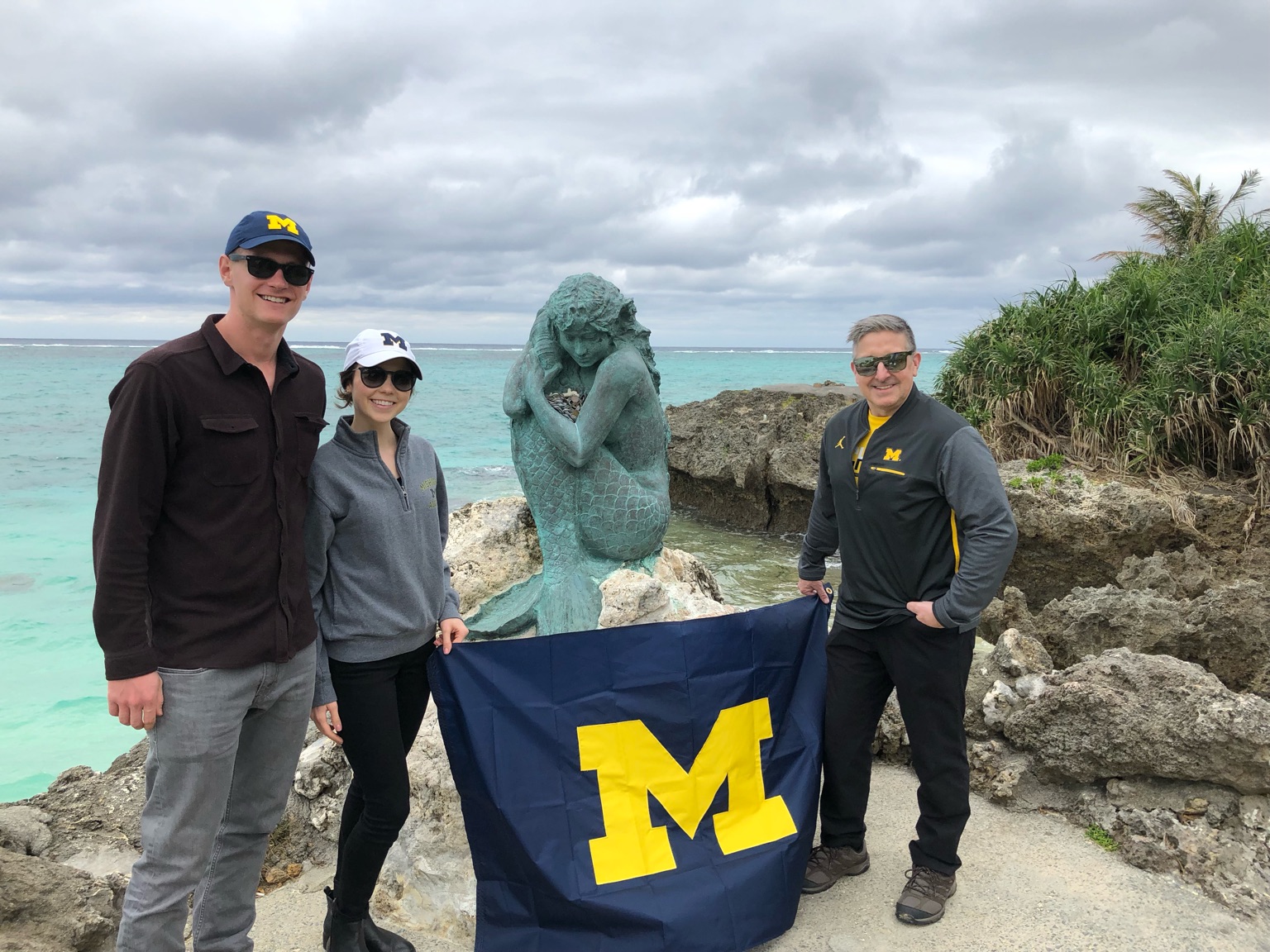 Chris Harkey, ’83, MSE’90, Paulina Singer, ’16, and Jacob Singer, ’16, waved the Michigan banner at the mermaid statue of Moon Beach on Okinawa Island, Japan.