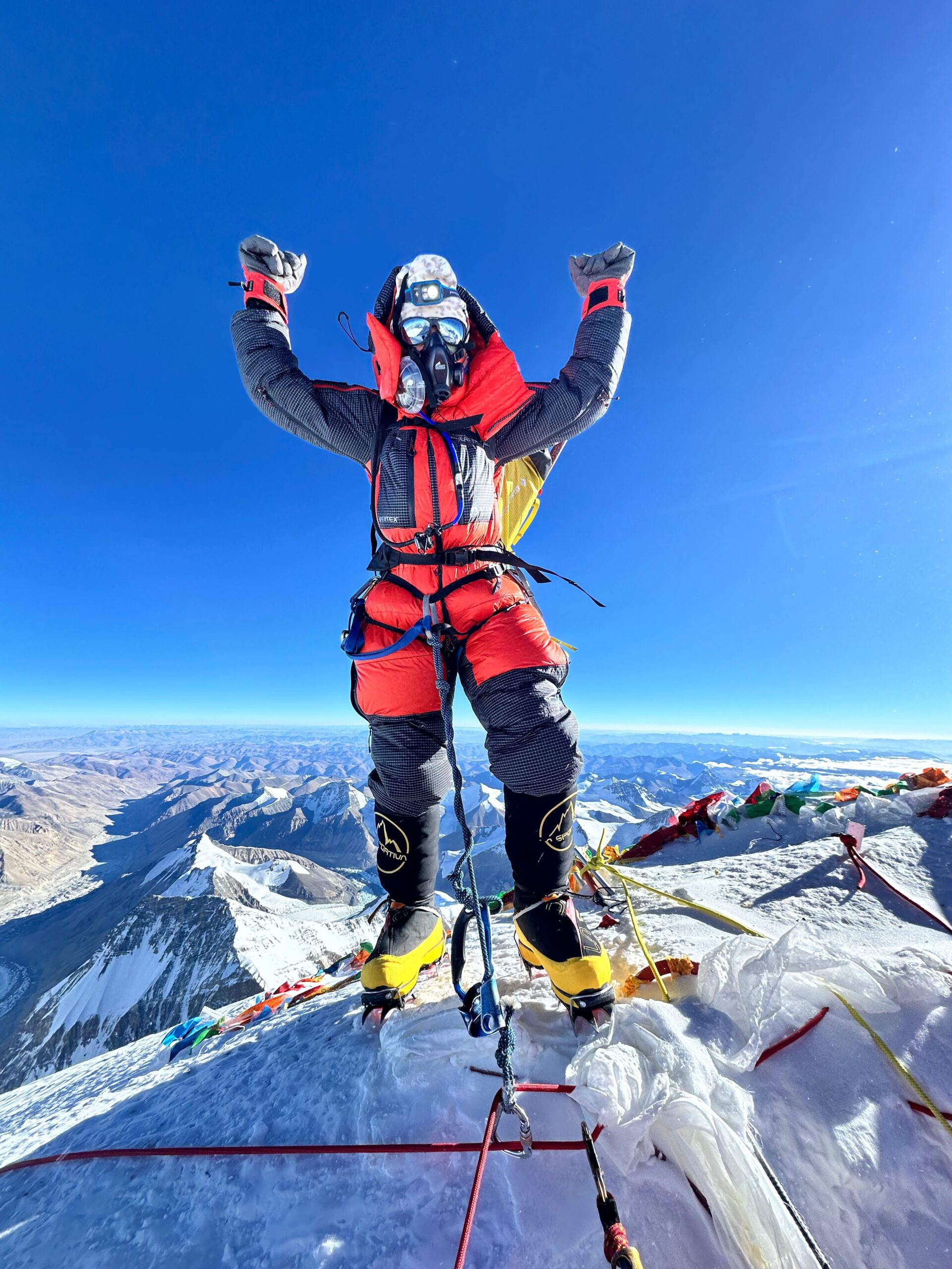 Myra Beaudoin Bertrand standing in her orange snow suit with her arms raised on top of Everest.