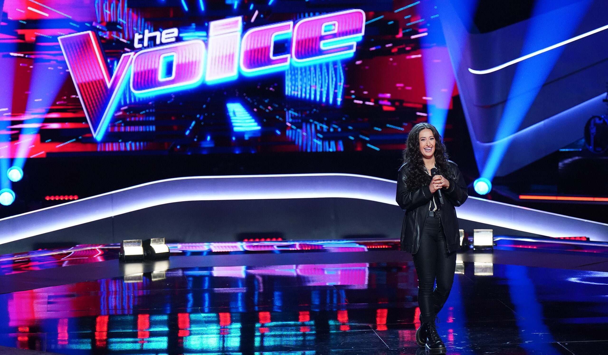 Frankie Torres, wearing all black, is smiling and holding a microphone on the right side of a stage with a large "The Voice" sign in the background.