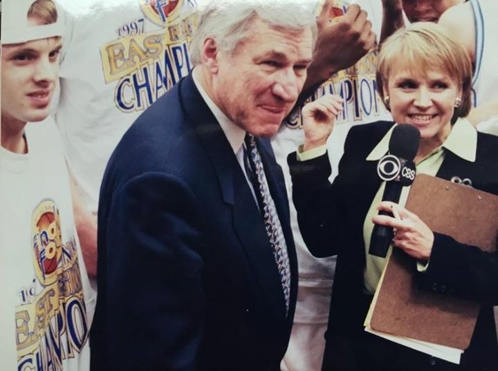 Joyce, right, holds a CBS microphone to her mouth and holds a clipboard in her right arm. Dean Smith, left, is in a blue blazer, white shirt, and tie. There are people behind them wearing 1997 Basketball National Championship shirts.