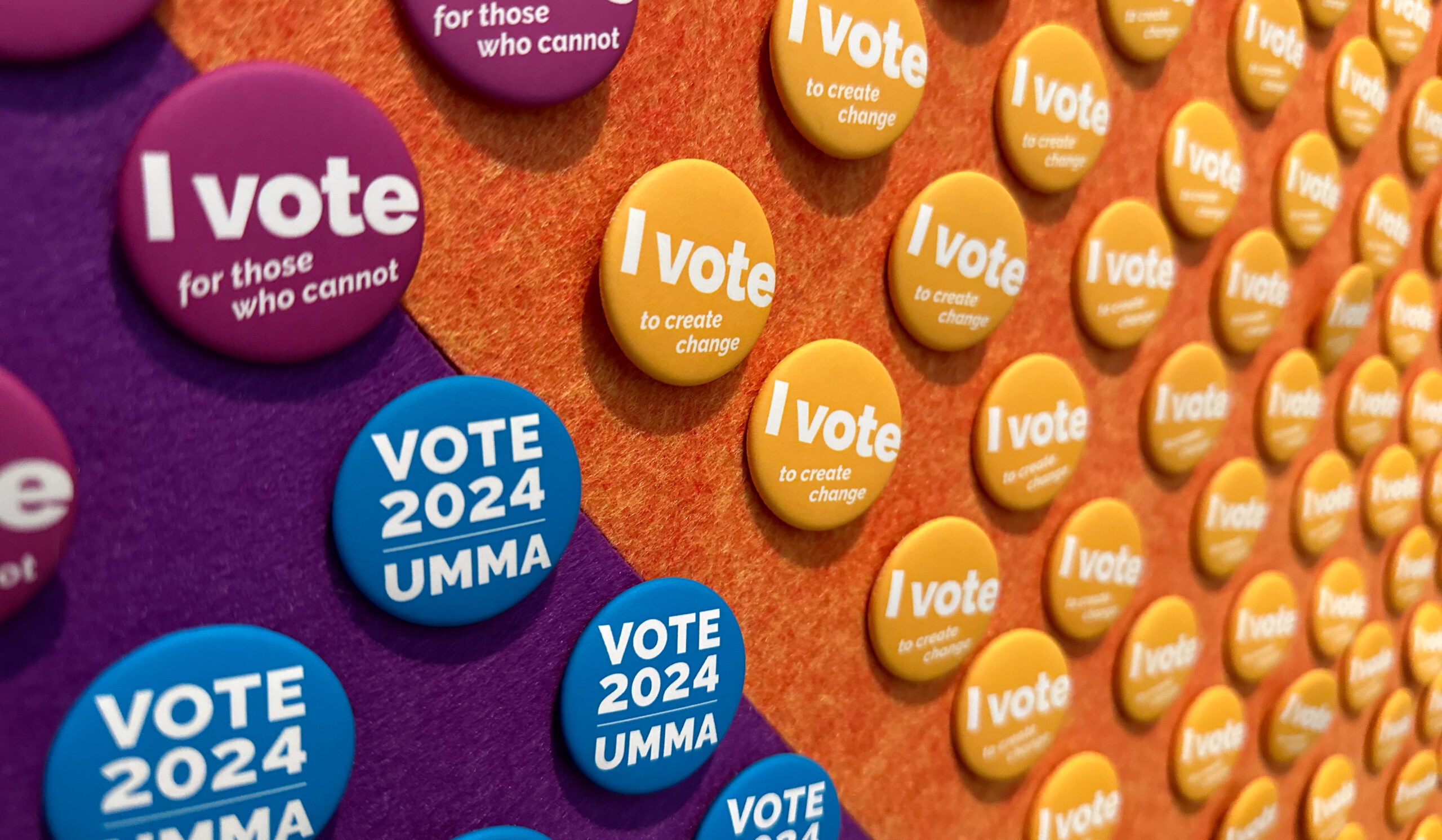 Round buttons are pinned to an orange and purple wall. The yellow one says "I vote to create change," the purple ones say "I vote for those who cannot," and the blue ones say "Vote 2024 UMMA."